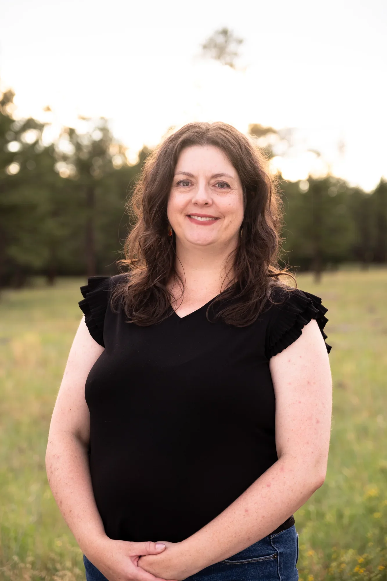 A woman with long, curly brown hair, smiling, standing outside in a field with trees in the background during sunset, wearing a black top with ruffled sleeves.