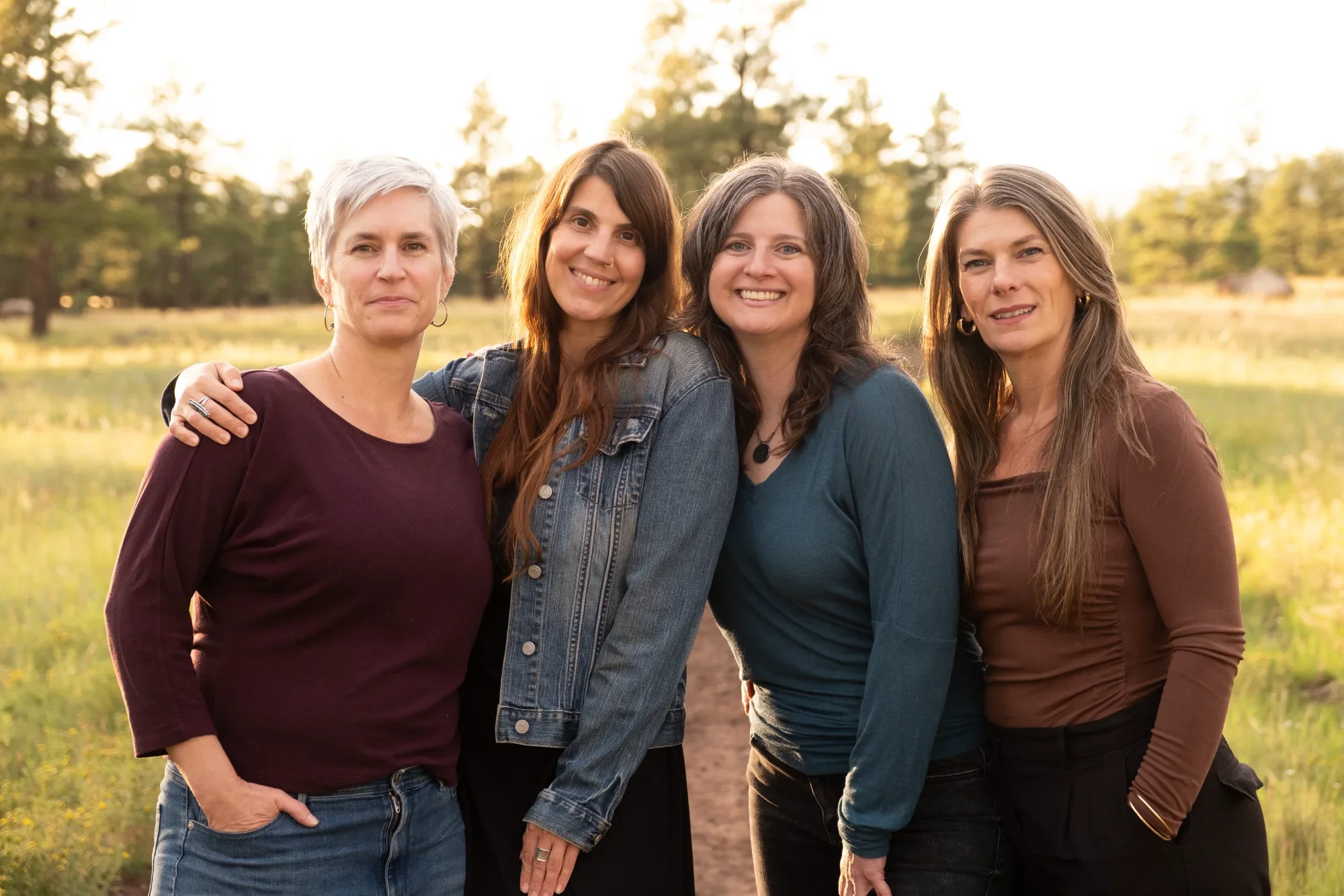 Four women standing close together outdoors during sunset, smiling at the camera. The background includes trees and a serene lake.