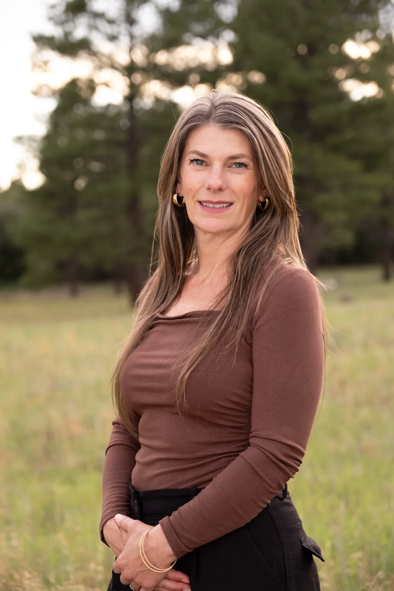 A woman with long brown hair, wearing a brown long-sleeve top and black pants, standing outdoors in a field with trees in the background. She is smiling and posing with her hands clasped in front of her.
