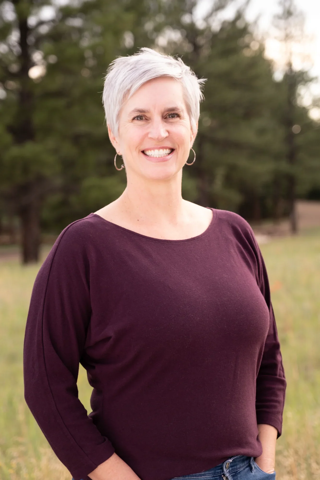 A woman with short gray hair smiling outdoors in a park with trees in the background, wearing a dark purple long-sleeve top and blue jeans.