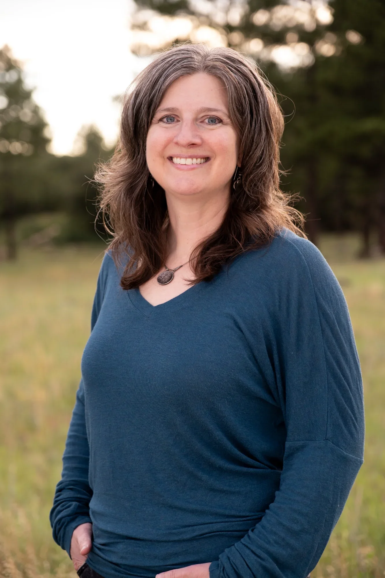 A smiling woman with shoulder-length brown hair and blue eyes, wearing a blue long-sleeve top and a necklace, standing outdoors in a grassy field with trees in the background at sunset.