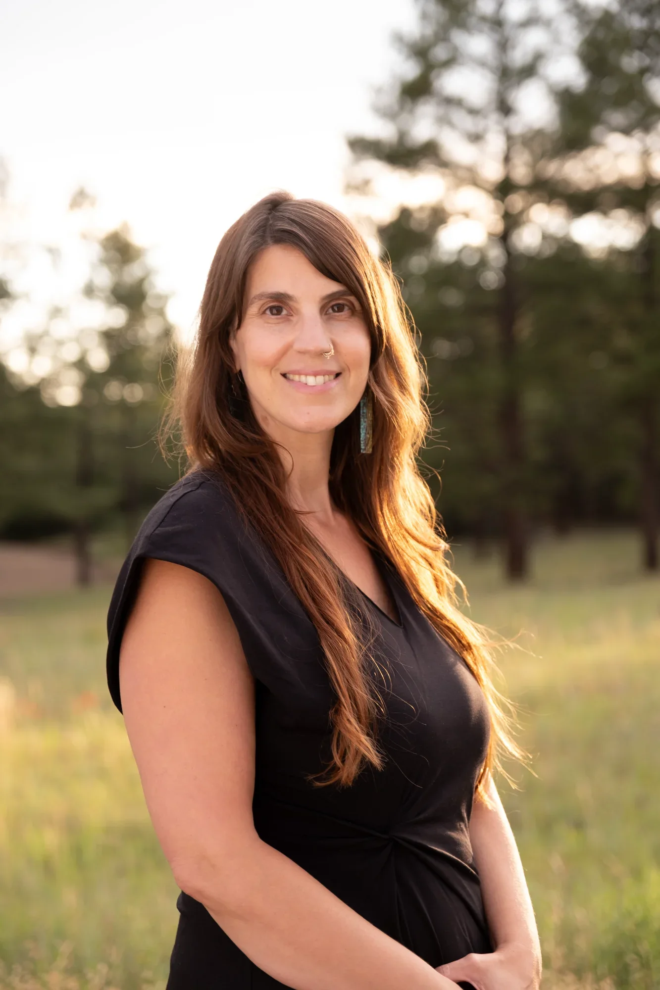A woman with long brown hair, wearing a black dress and turquoise earrings, standing outdoors during sunset with trees in the background.