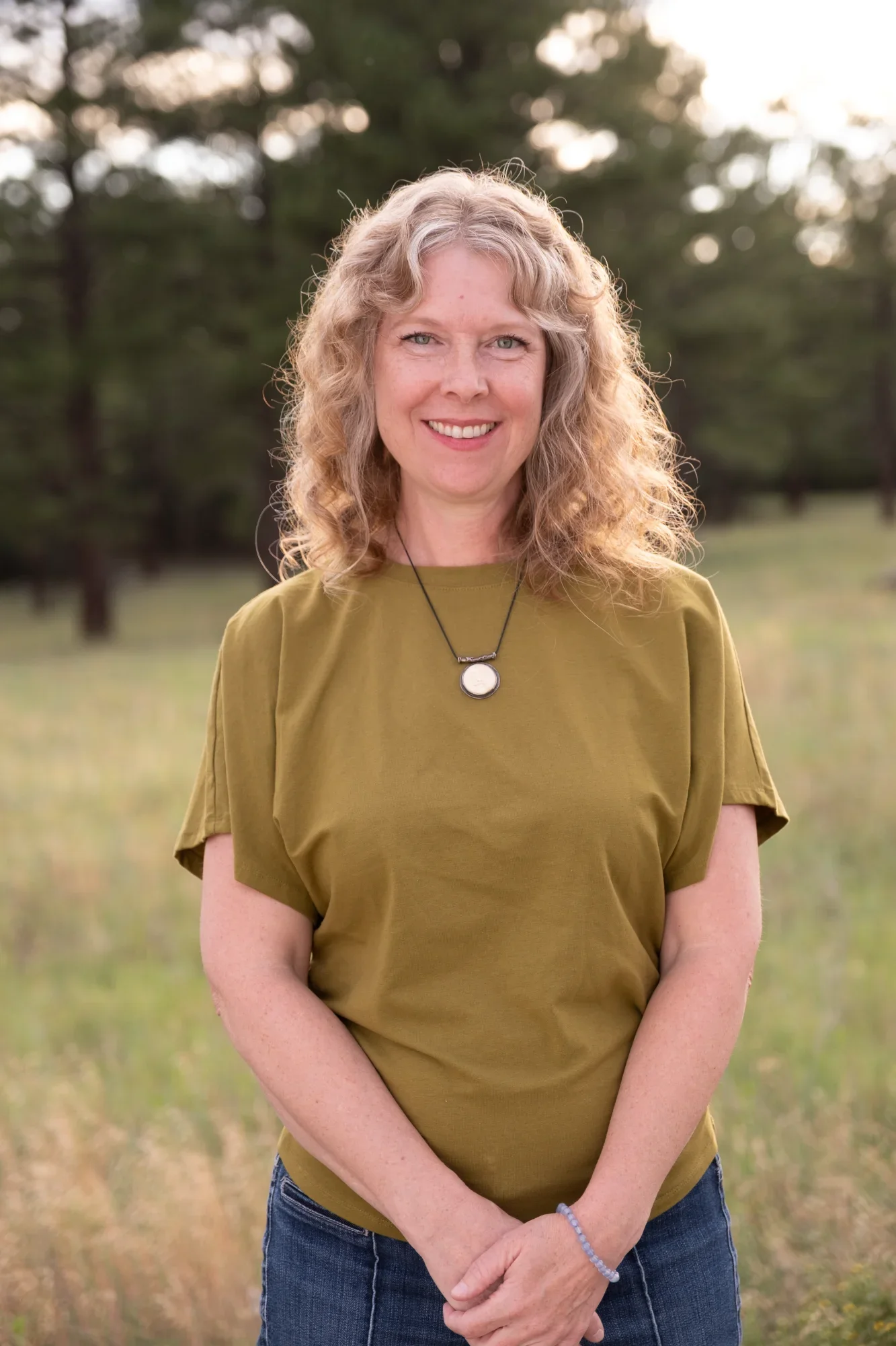 Portrait of a woman with curly blonde hair smiling outdoors, wearing a green t-shirt and jeans, with trees in the background at sunset.