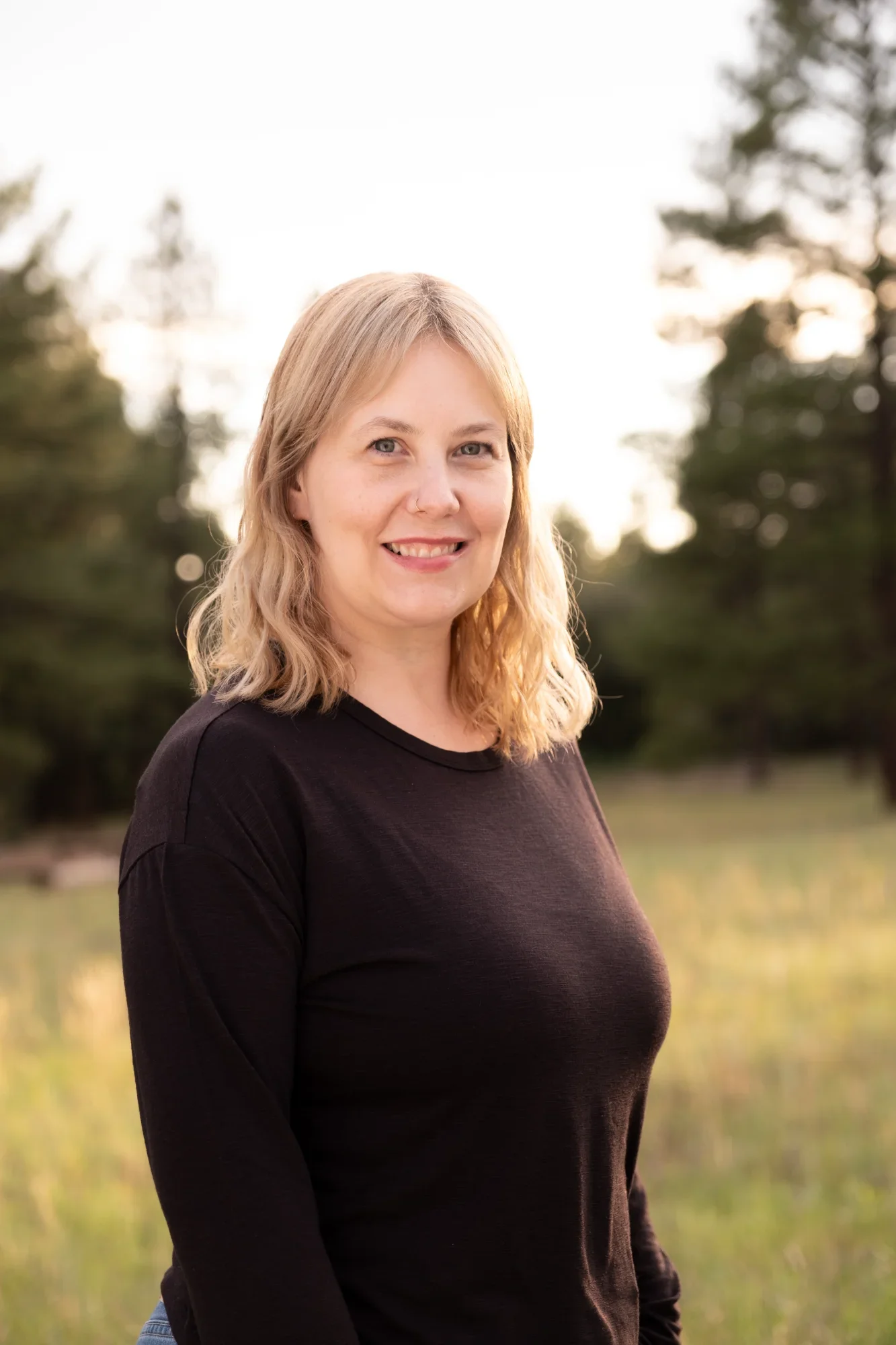A woman with blonde wavy hair smiling outdoors, wearing a black long sleeve shirt, with a blurred background of trees and sky.
