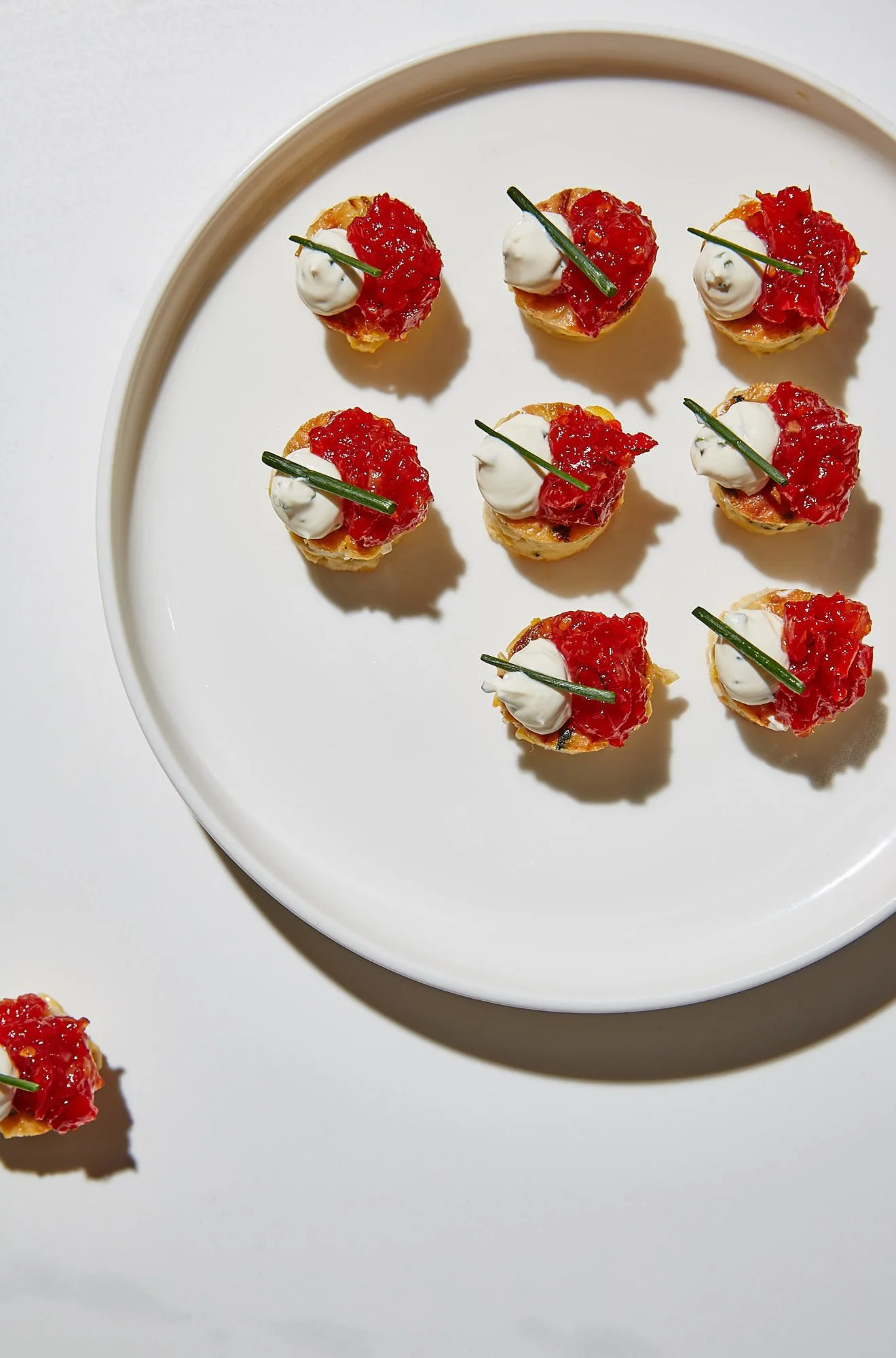 A white plate with nine small hors d'oeuvres topped with red sauce, a dollop of cream, and a chive garnished.