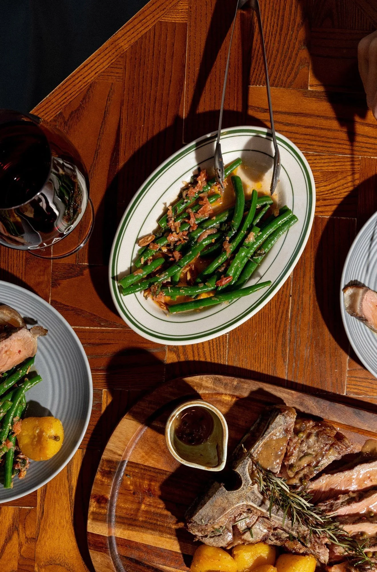Steak resting on a wooden cutting board with rosemary, potatoes, and a small dish of sauce, surrounded by other dishes including green beans with bacon, a glass of red wine, and other plated foods on a wooden table.