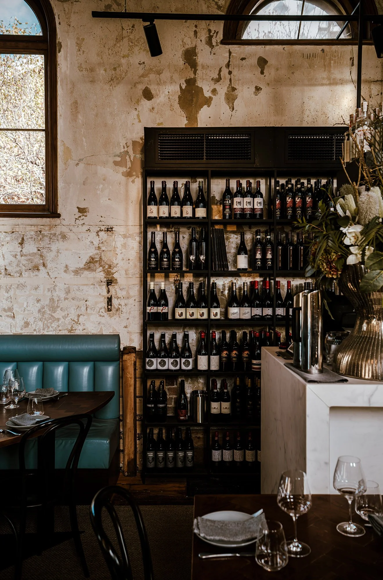 A wine display shelf filled with bottles of wine, located in a stylish, rustic restaurant with an exposed brick and peeling plaster wall, large windows letting in natural light, and a cozy dining area with place settings and wine glasses on tables.