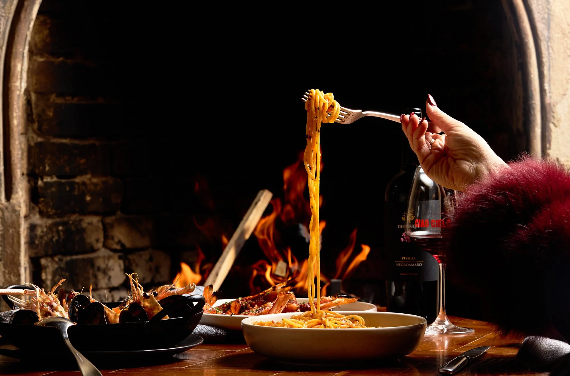 Hand holding a fork with spaghetti pasta above a bowl of spaghetti, in front of a brick fireplace with flames. A glass of red wine is on the table.