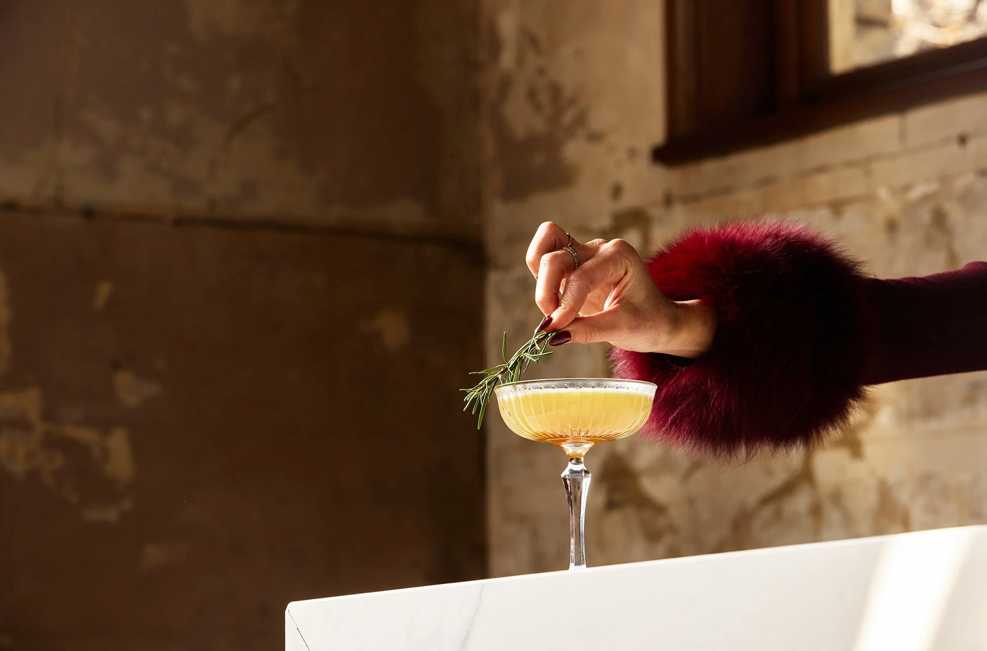 A hand holding a sprig of rosemary above a coupe glass filled with a yellow cocktail, with a brick wall and a window in the background.