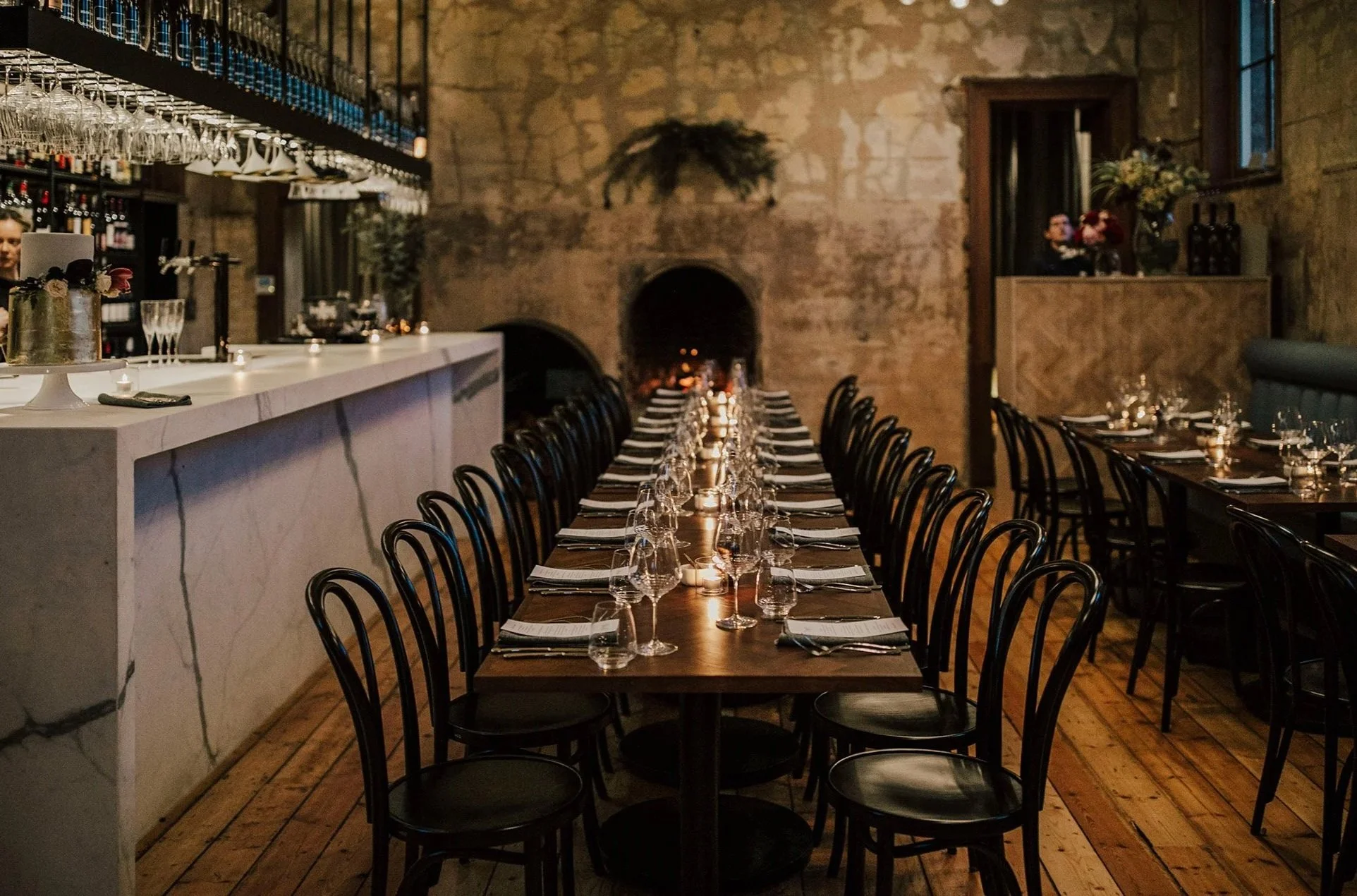 Empty restaurant with a long dining table set with glasses, plates, and napkins, candles, and a marble bar counter with baristas preparing drinks. Exposed brick walls and large windows.