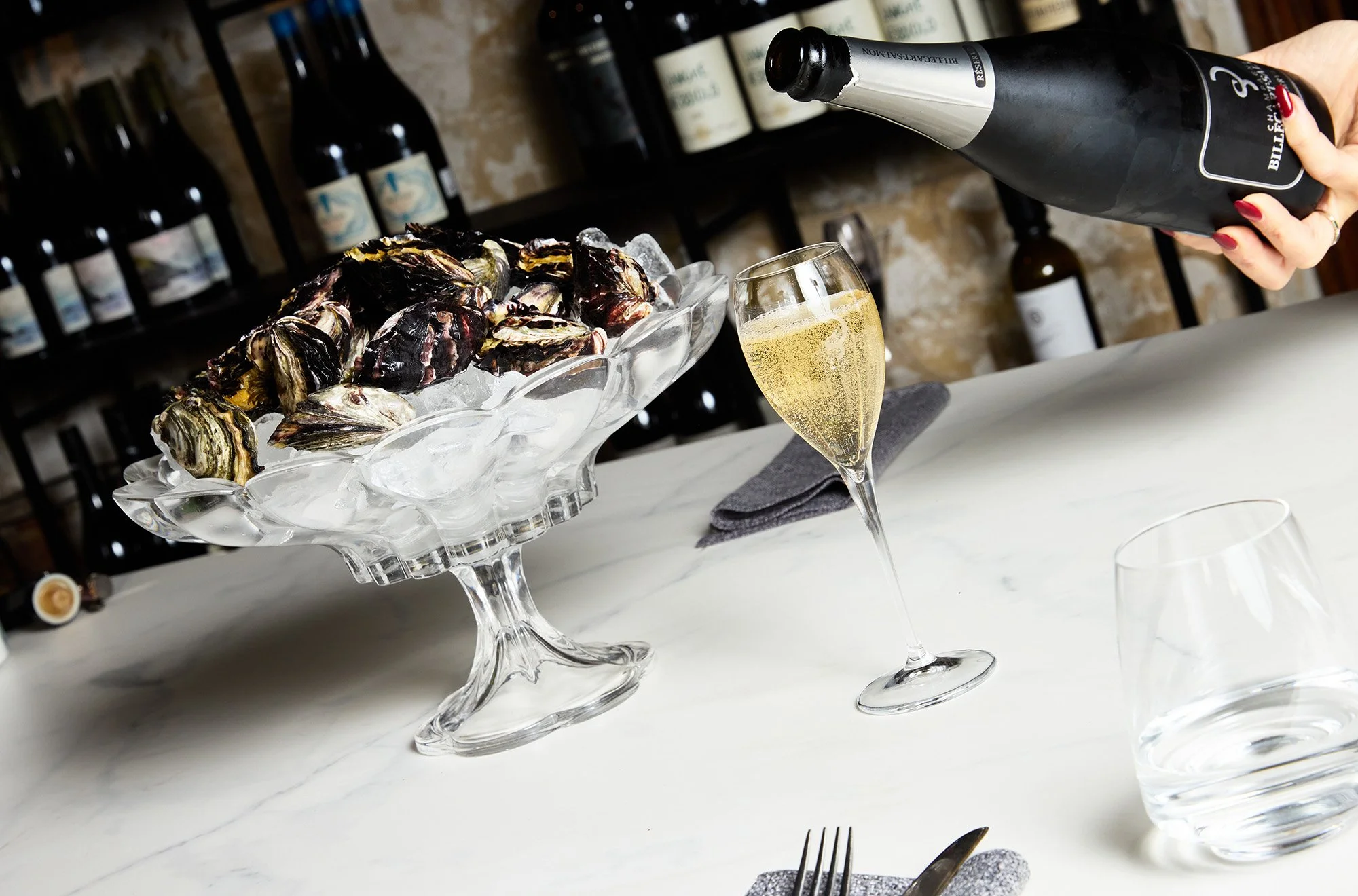 A hand pouring champagne into a flute glass near a glass dish filled with oysters on ice at a dining table.
