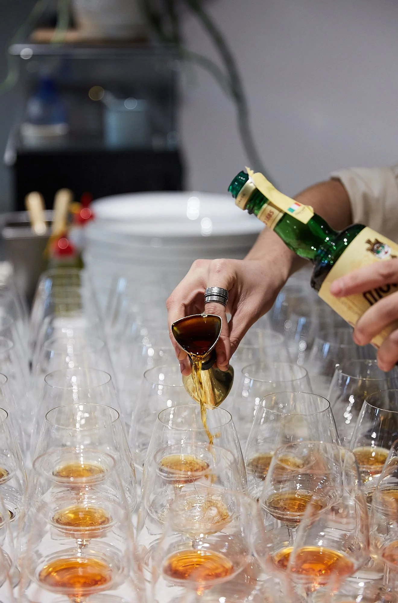 Person pouring a drink into a glass at a bar or event, with multiple glasses arranged in front.
