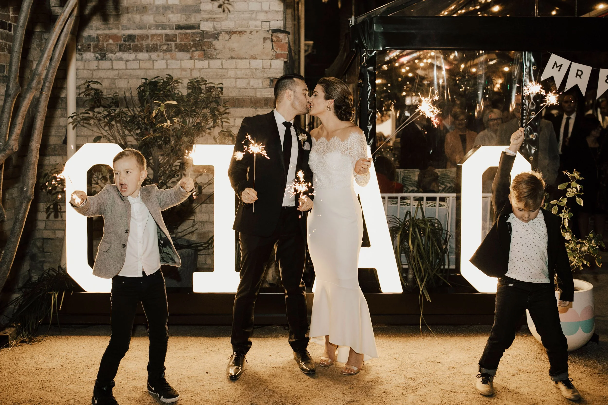 A wedding scene at night showing a bride and groom kissing, surrounded by children holding sparklers and celebrating. The background features a brick wall and festive decorations.