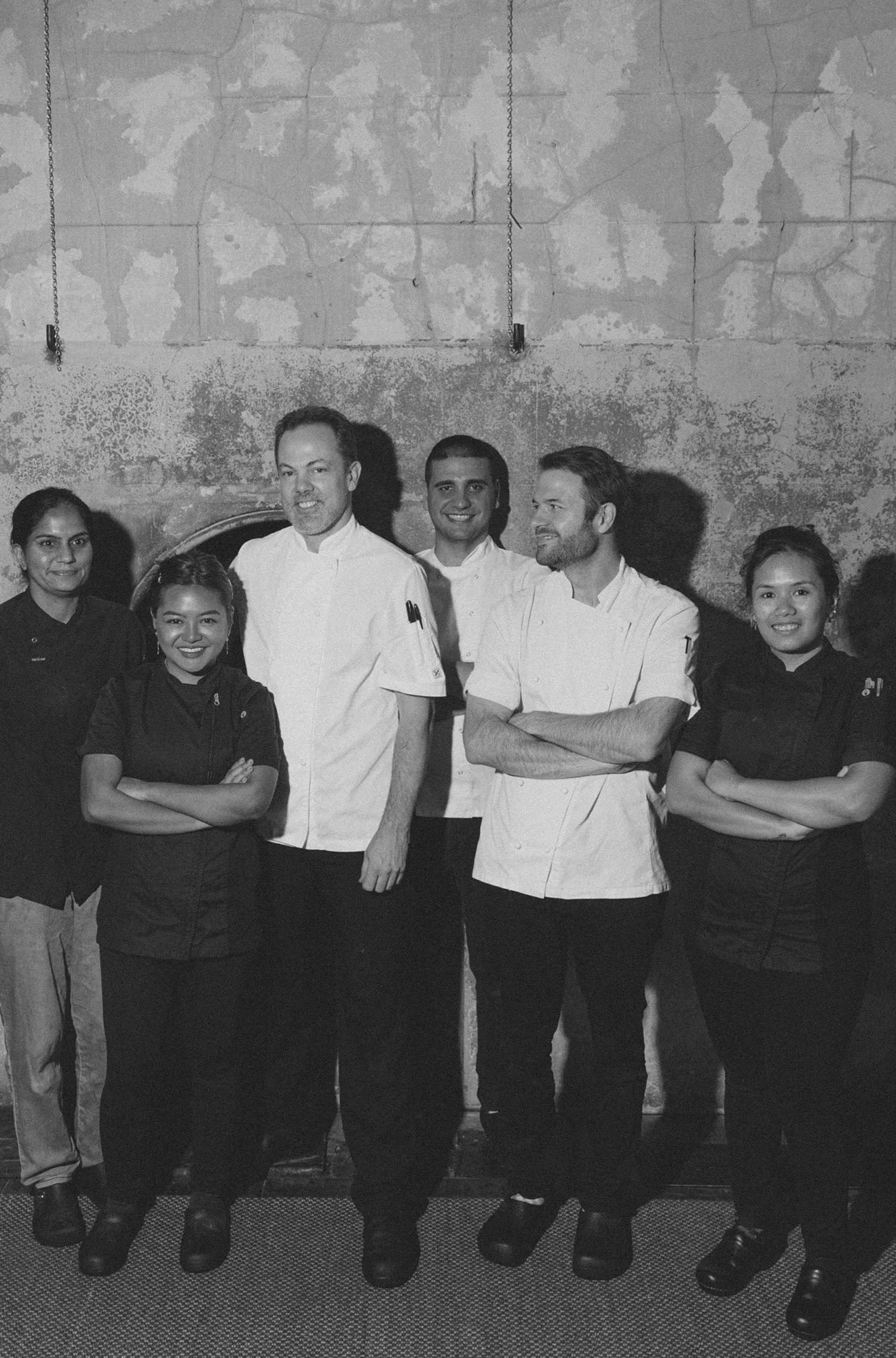 Group of six chefs and staff standing together, smiling, in a kitchen or restaurant setting with a textured wall background.
