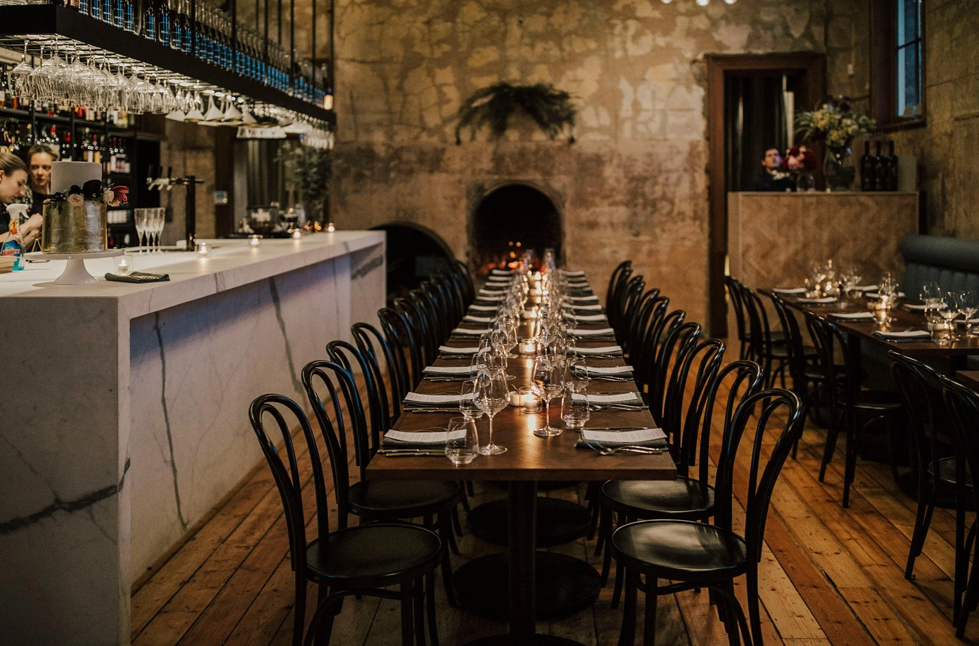 Empty restaurant with a long dining table set with glasses, plates, and napkins, candles, and a marble bar counter with baristas preparing drinks. Exposed brick walls and large windows.