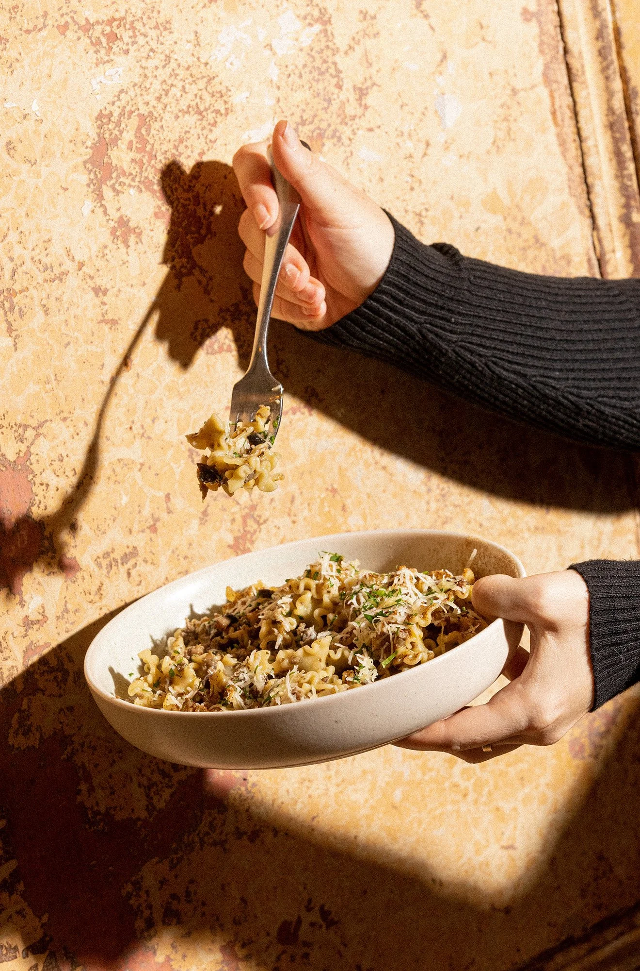 Person holding a white bowl filled with cooked pasta, ground meat, and garnish, while using a fork to serve the dish, against a textured beige wall.