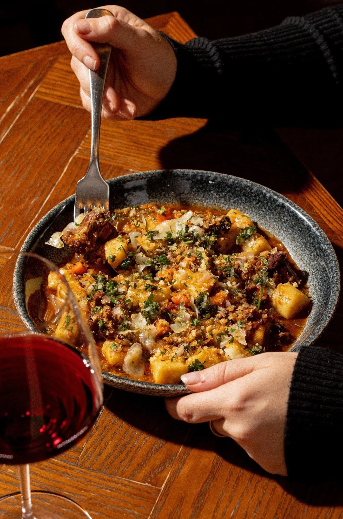 A person in a black sweater is holding a fork and a bowl of hearty stew with potatoes, vegetables, and herbs. There is a glass of red wine and a wooden table in the background.