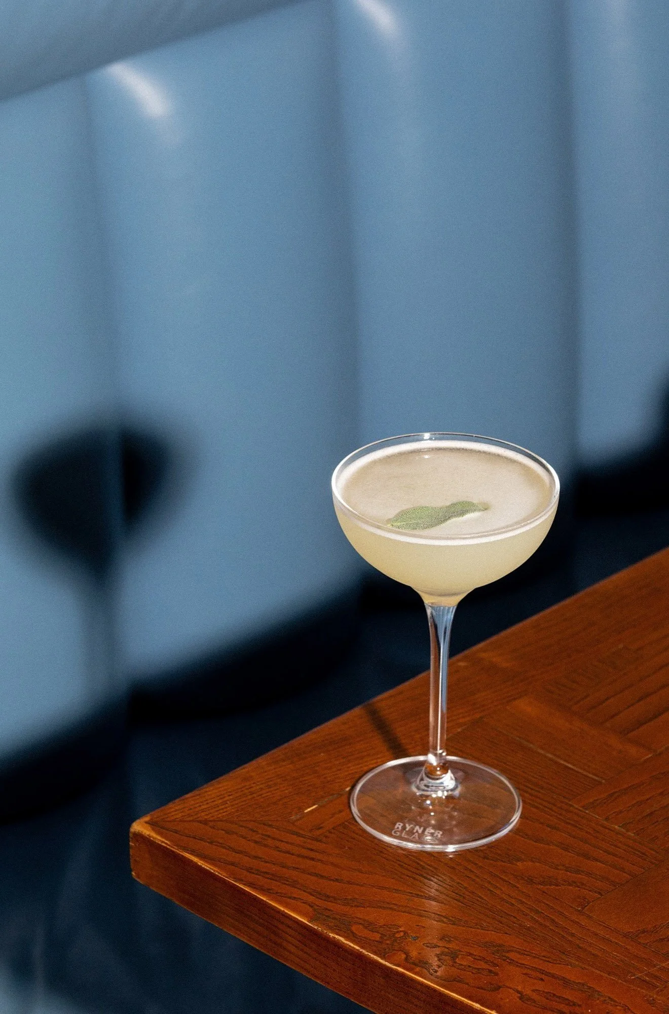 A cocktail glass with a pale yellow drink garnished with a sage leaf, placed on a wooden surface.