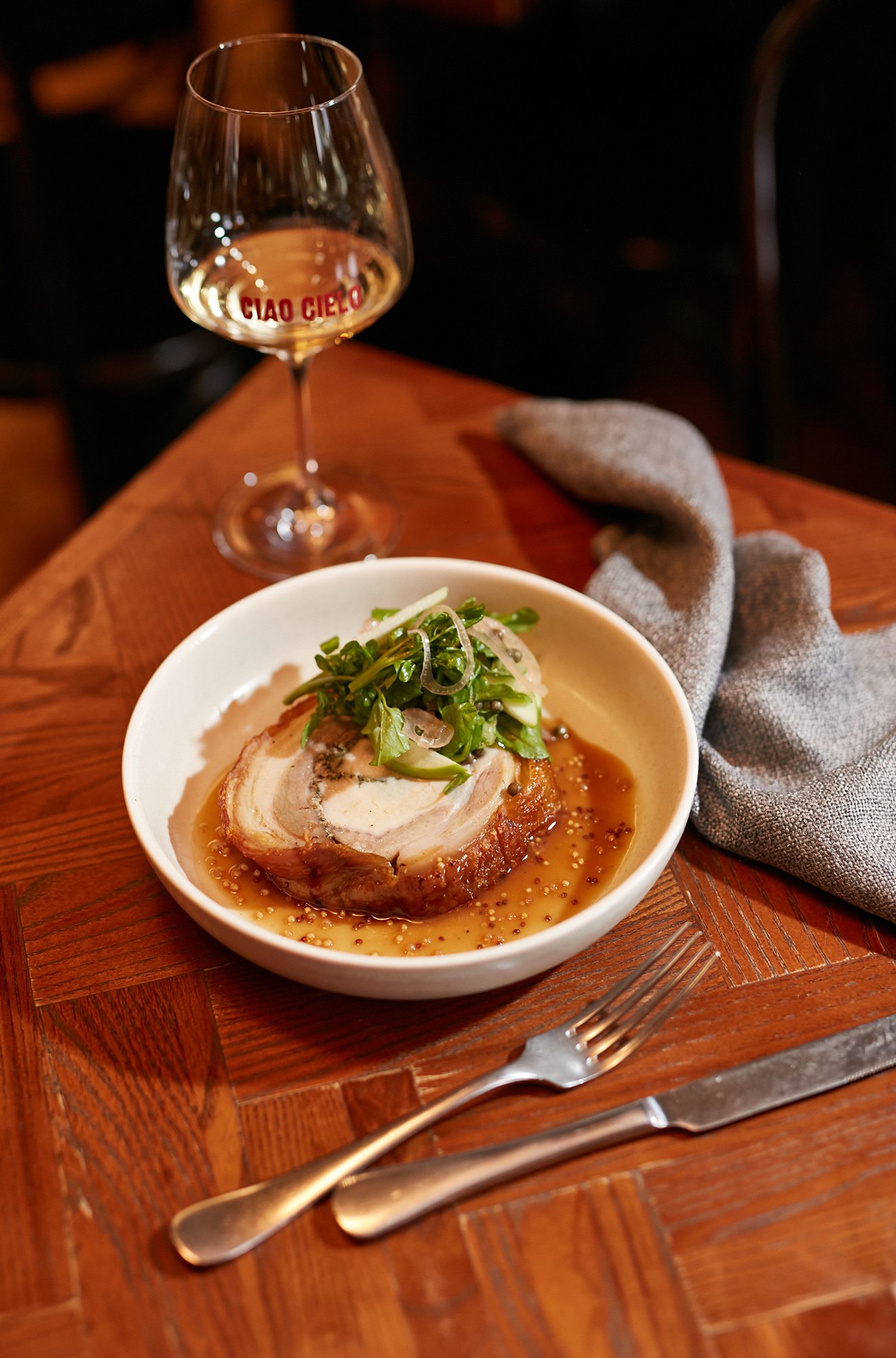 A bowl of roasted pork stuffed with herbs topped with arugula, served with mustard seeds and sauce on a wooden table. In the background, a glass of white wine with the words 'CIAO CIELO' printed on it, and a gray linen napkin nearby.