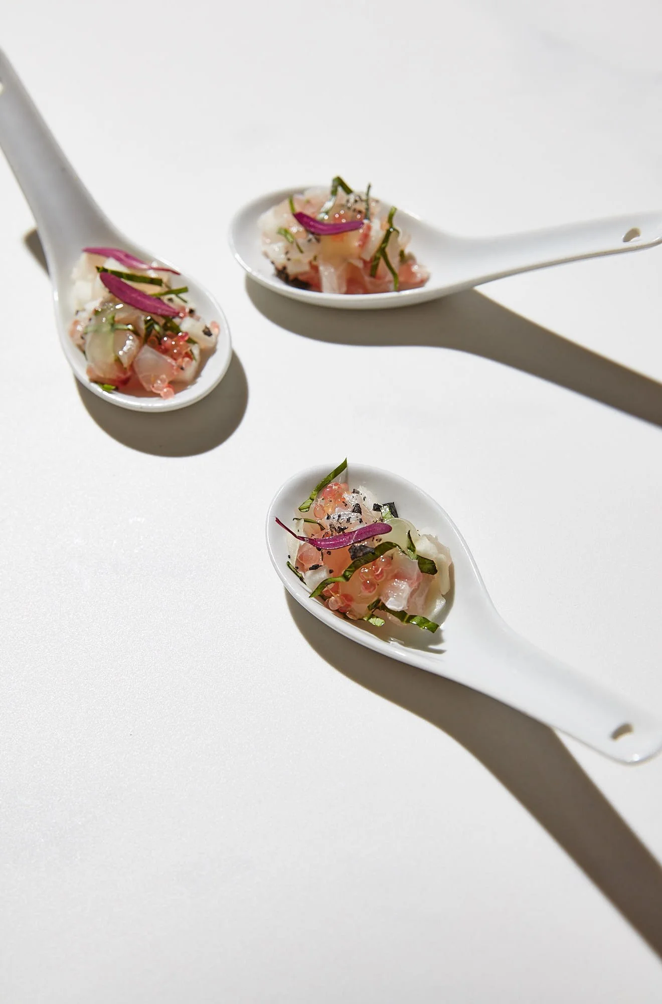 Three white porcelain spoons filled with chopped raw fish, garnished with thinly sliced red onions, green herbs, and black sesame seeds, displayed on a white surface.