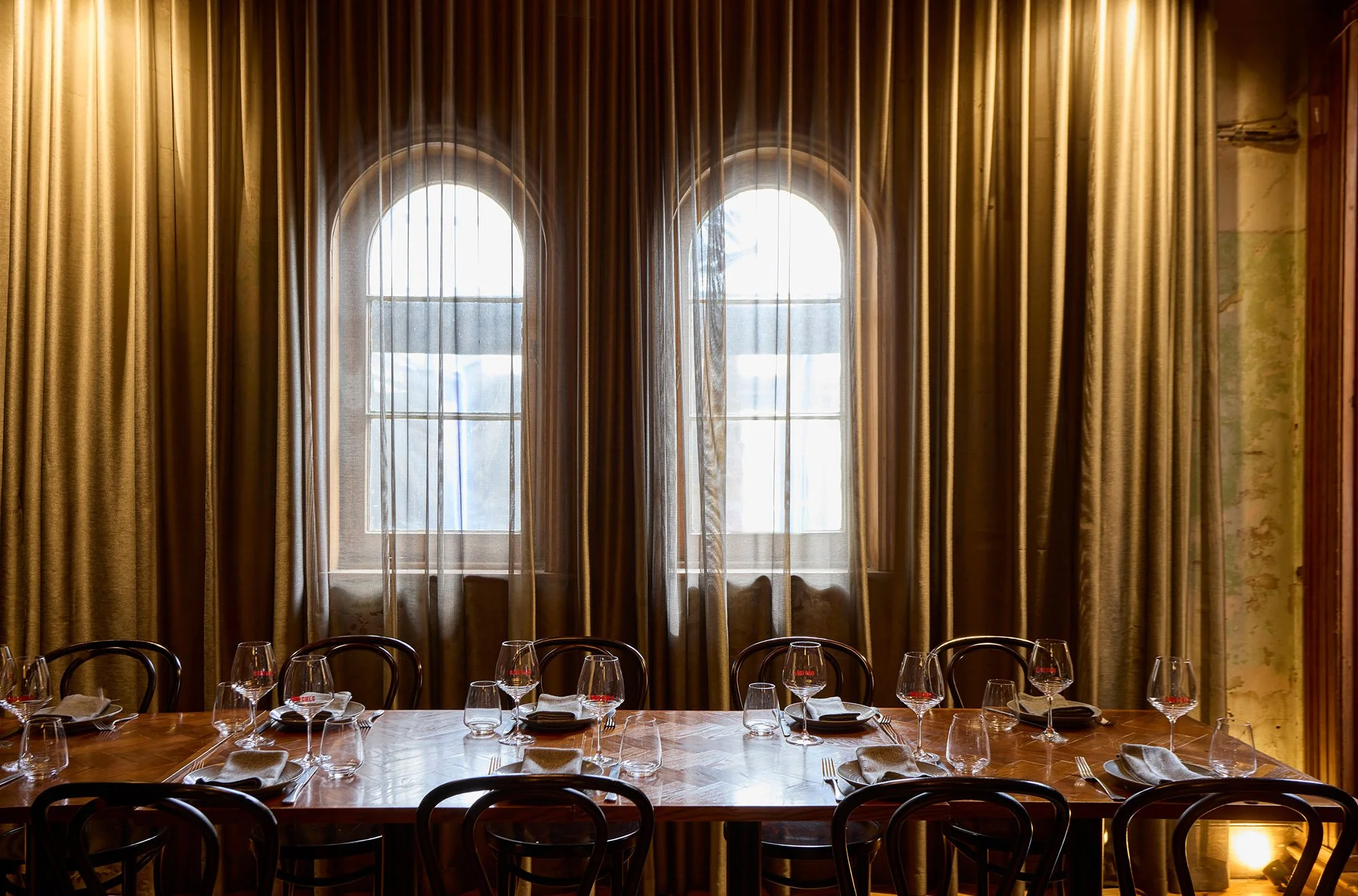 Elegant dining room with a large, polished wooden table, multiple wine glasses, and napkins set for a meal, framed by tall curtains and two arched windows in the background.