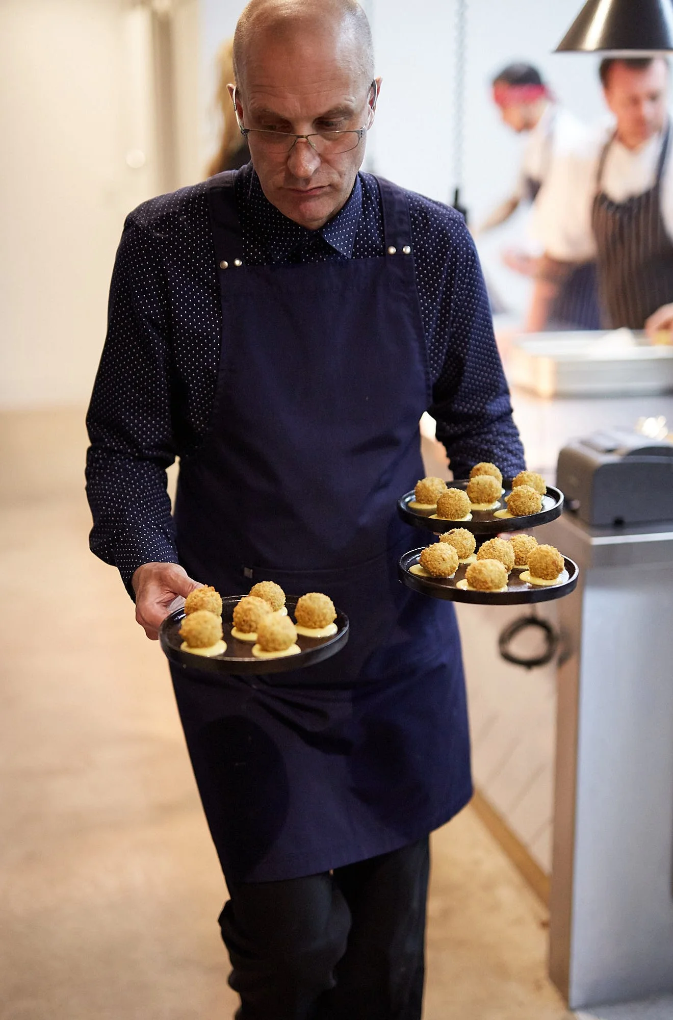 A waiter with glasses wearing a navy apron serving two trays of appetizers with breaded balls in a restaurant kitchen.