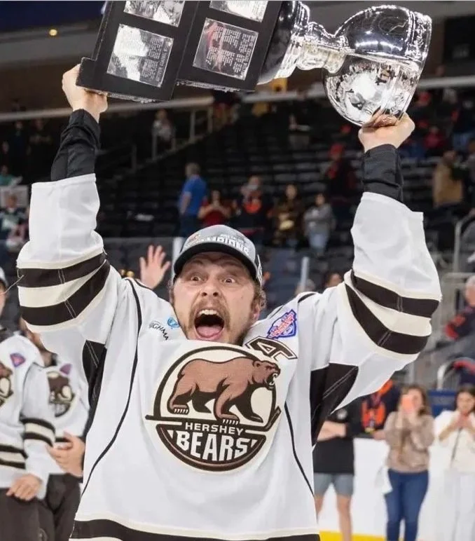 A hockey player wearing a Hershey Bears jersey celebrating with arms raised after winning a championship, holding a large trophy above his head, in an ice rink with fans and other players in the background.