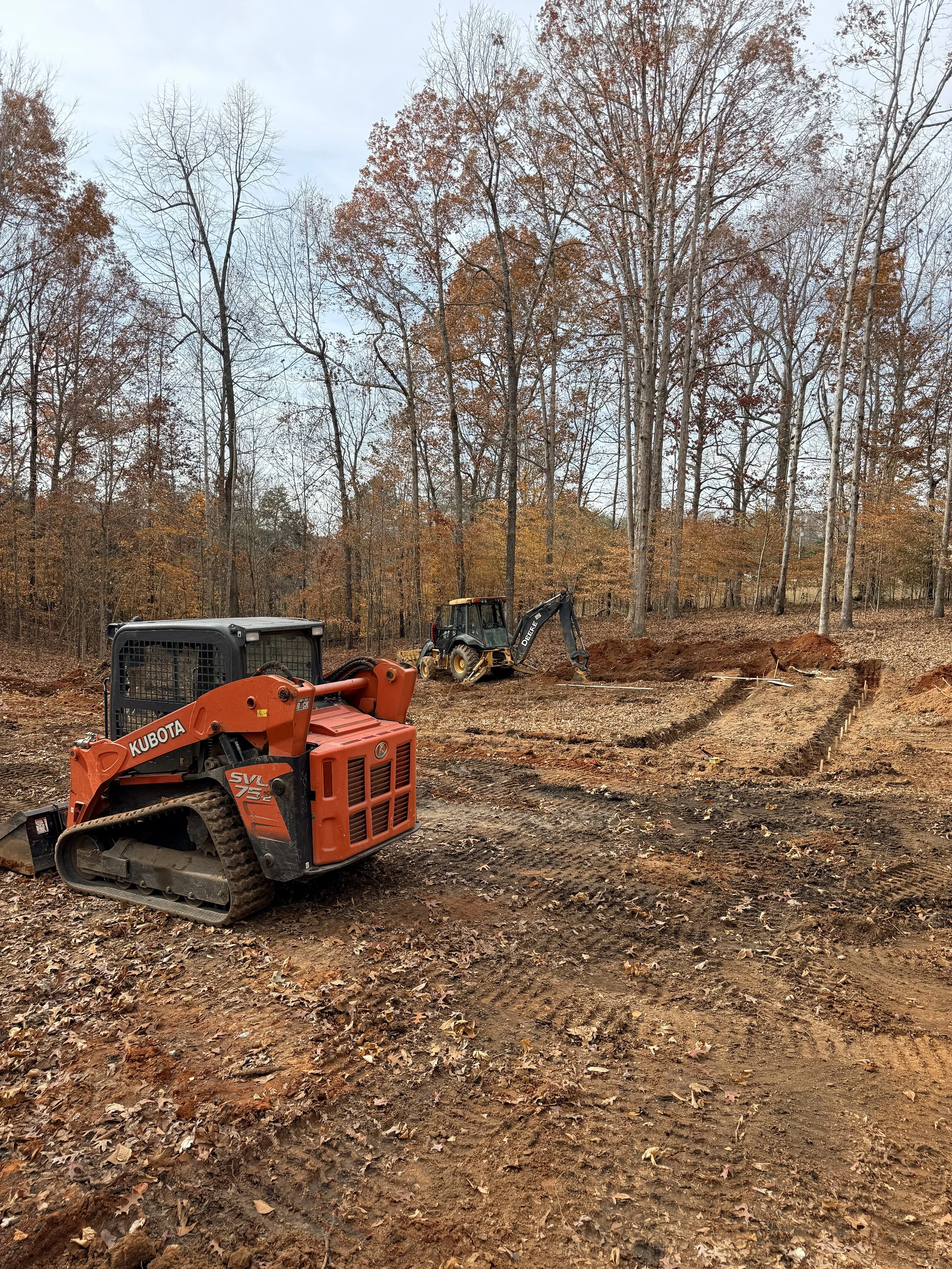 Construction site in a wooded area with excavated soil, a Kubota compact excavator, and a Deere backhoe loader working amidst fallen leaves and surrounded by tall trees with autumn foliage.