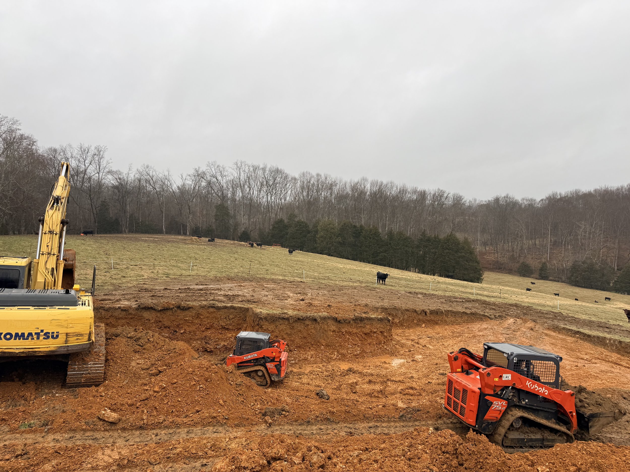 Construction site with heavy machinery, including a yellow excavator and small orange machines, digging into the ground. In the background, there are fields with cows and a line of leafless trees under overcast skies.