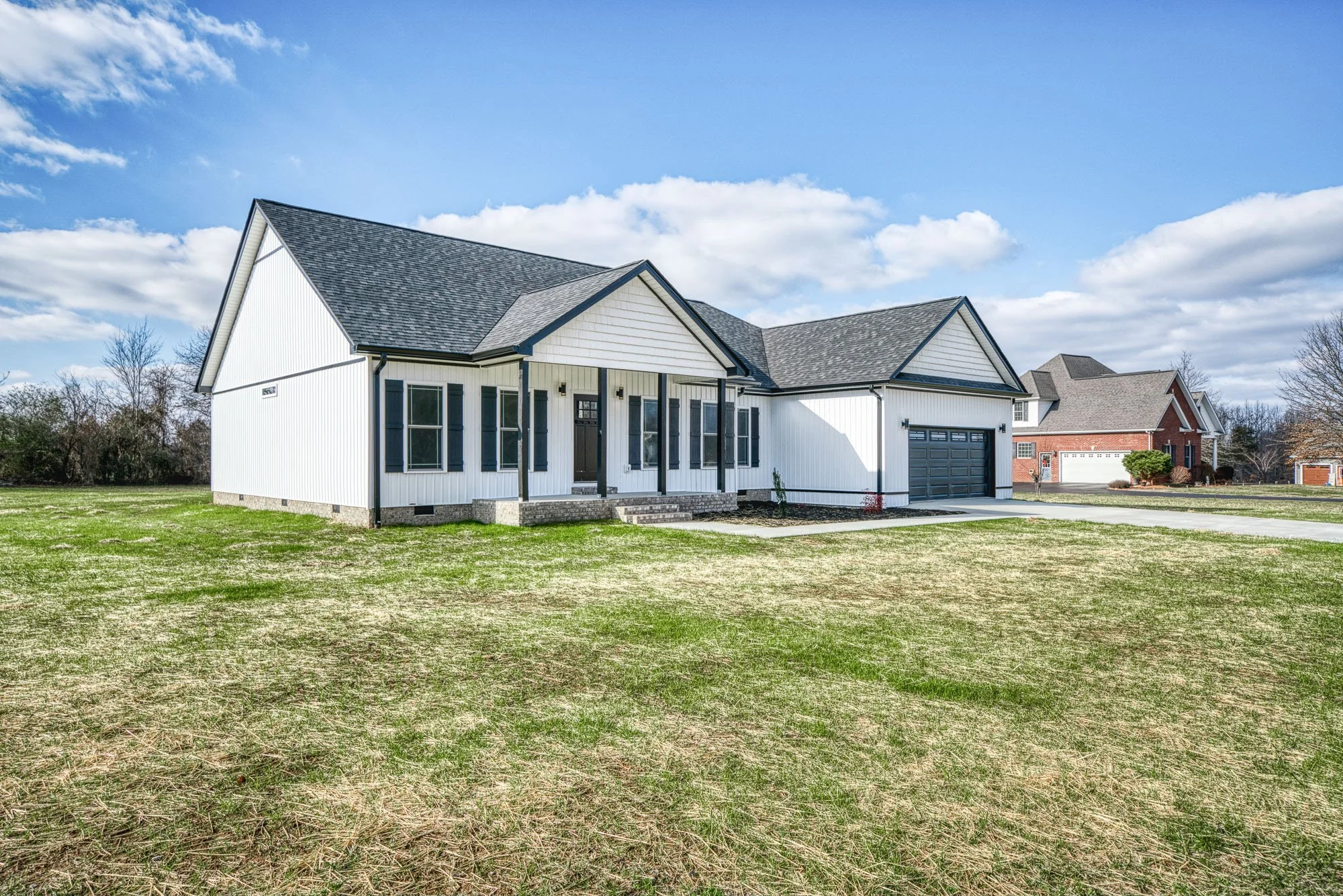 Front view of a modern white house with black shutters and a dark gray garage door, set on a lawn with a partly cloudy sky in the background.