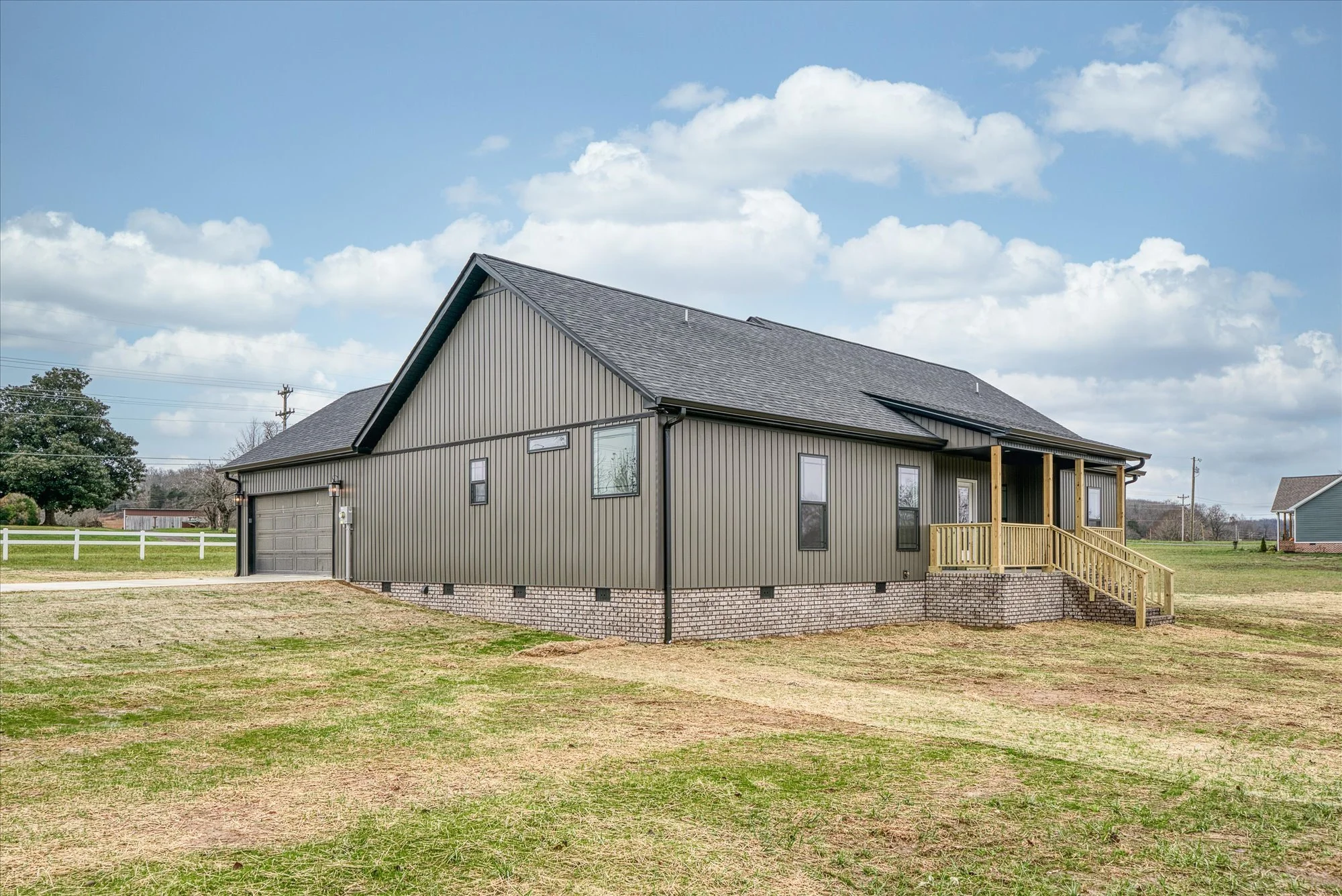 A modern two-story house with gray vertical siding, a dark gray roof, and a brick foundation. There is a small porch with a wooden railing and stairs. The house is situated in a grassy yard with a white fence and trees in the background under a partl