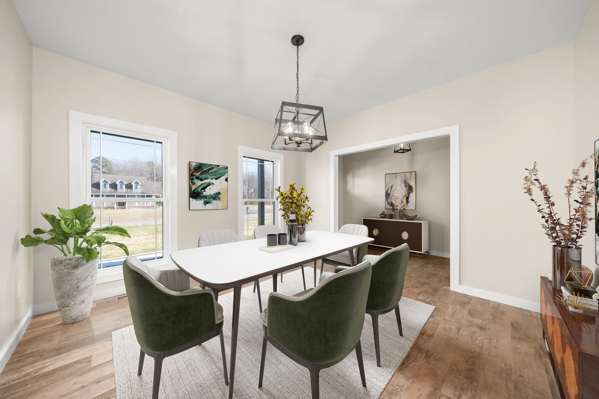 A dining room with a white table and six green velvet chairs. There are two windows letting in natural light, with abstract art on the walls. A modern black chandelier hangs above the table. The room has wooden floors, potted plants, and decorative a