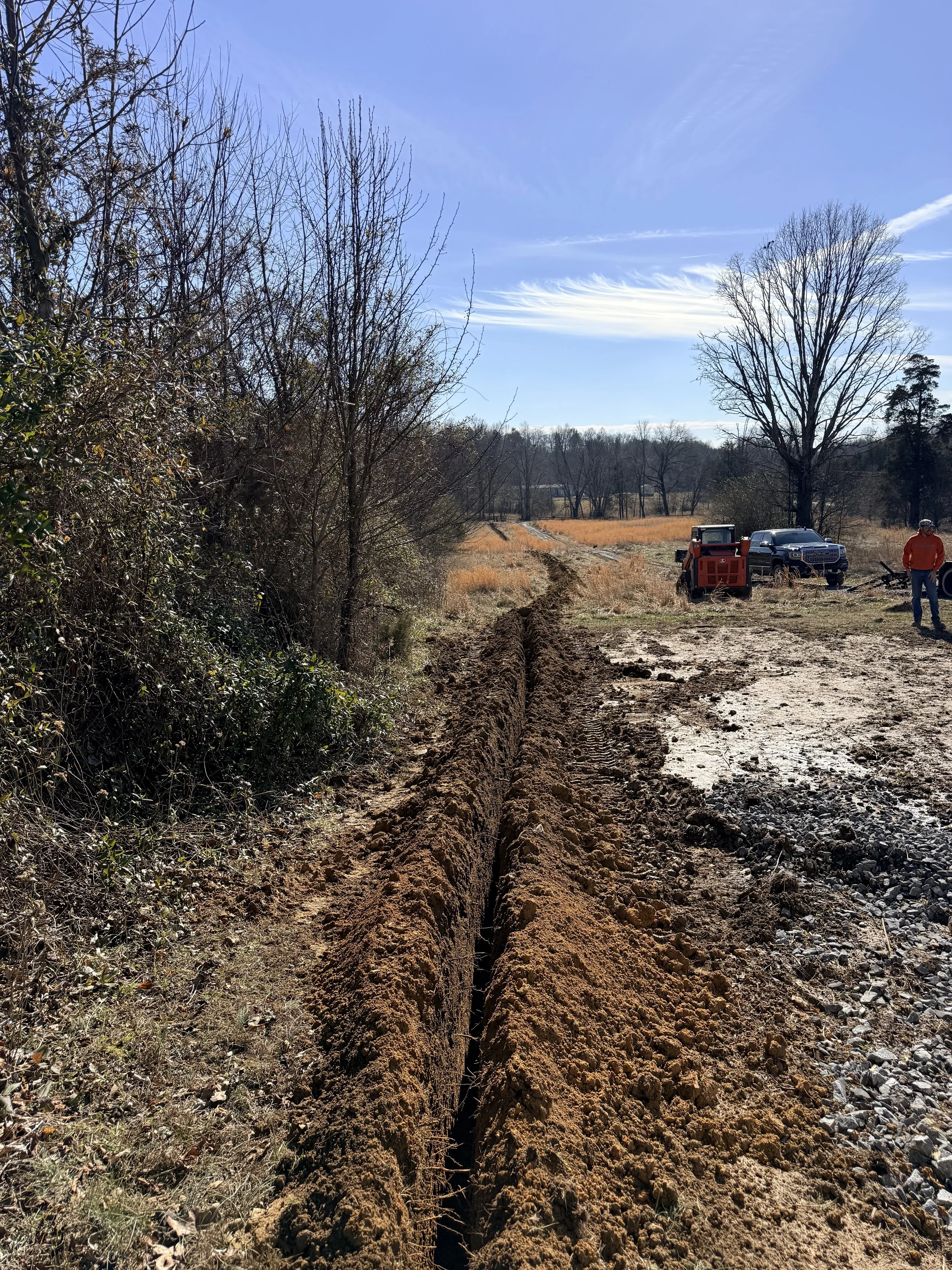 A dirt road preparing for utilities installation in an open rural area on a clear, sunny day with some clouds and leafless trees.