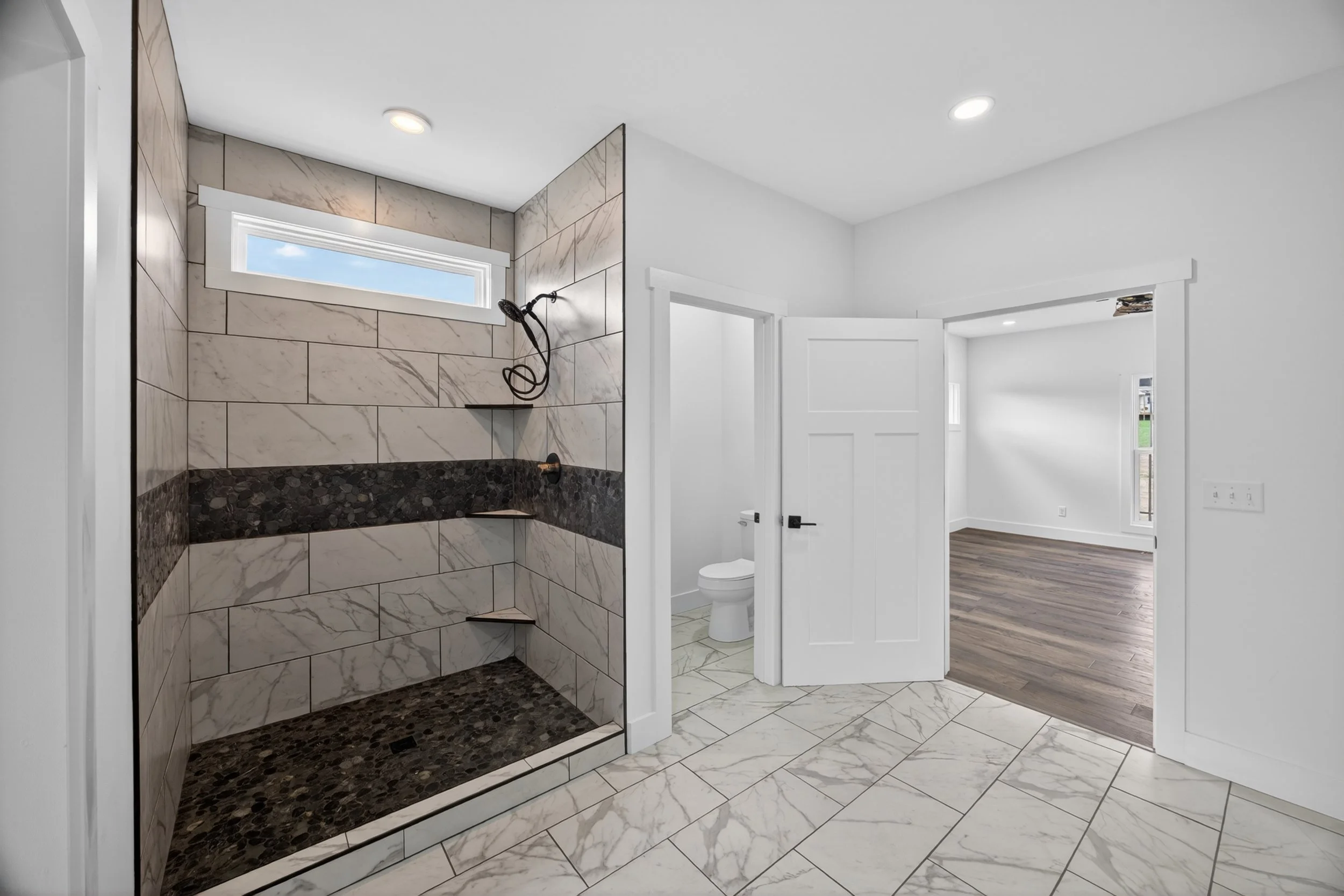 Modern bathroom with marble tile on floor and walls, featuring a walk-in shower with dark pebble stone at the bottom, a large window, and adjacent toilet. The room has white walls and ceiling with recessed lighting, and an open door leads to a bright