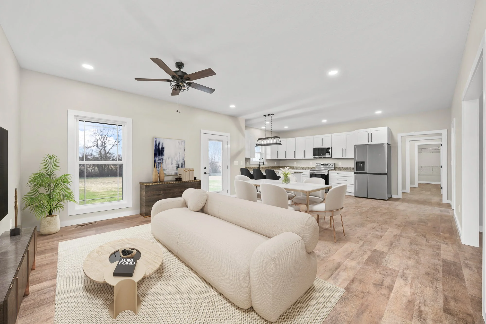 Open-concept living room and kitchen with white walls, light wood flooring, a ceiling fan, and recessed lighting. The living room has a beige sofa, a round coffee table, a potted plant, and a window showing a tree outside. The kitchen has white cabinets, stainless steel appliances, and a dining table with white chairs.
