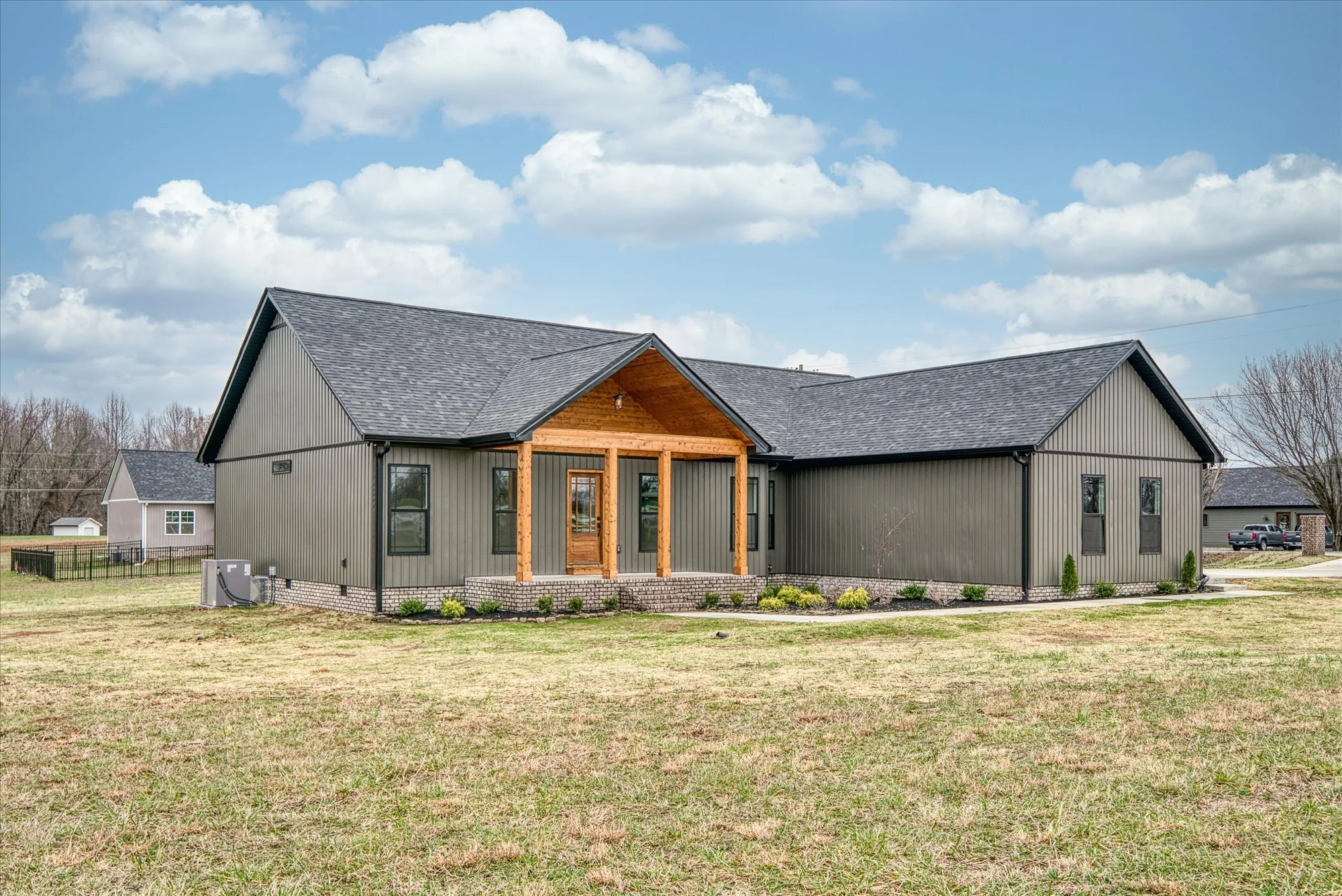New house with gray siding, black trim, and a gabled roof, located in a neighborhood with other houses, under a partly cloudy sky.