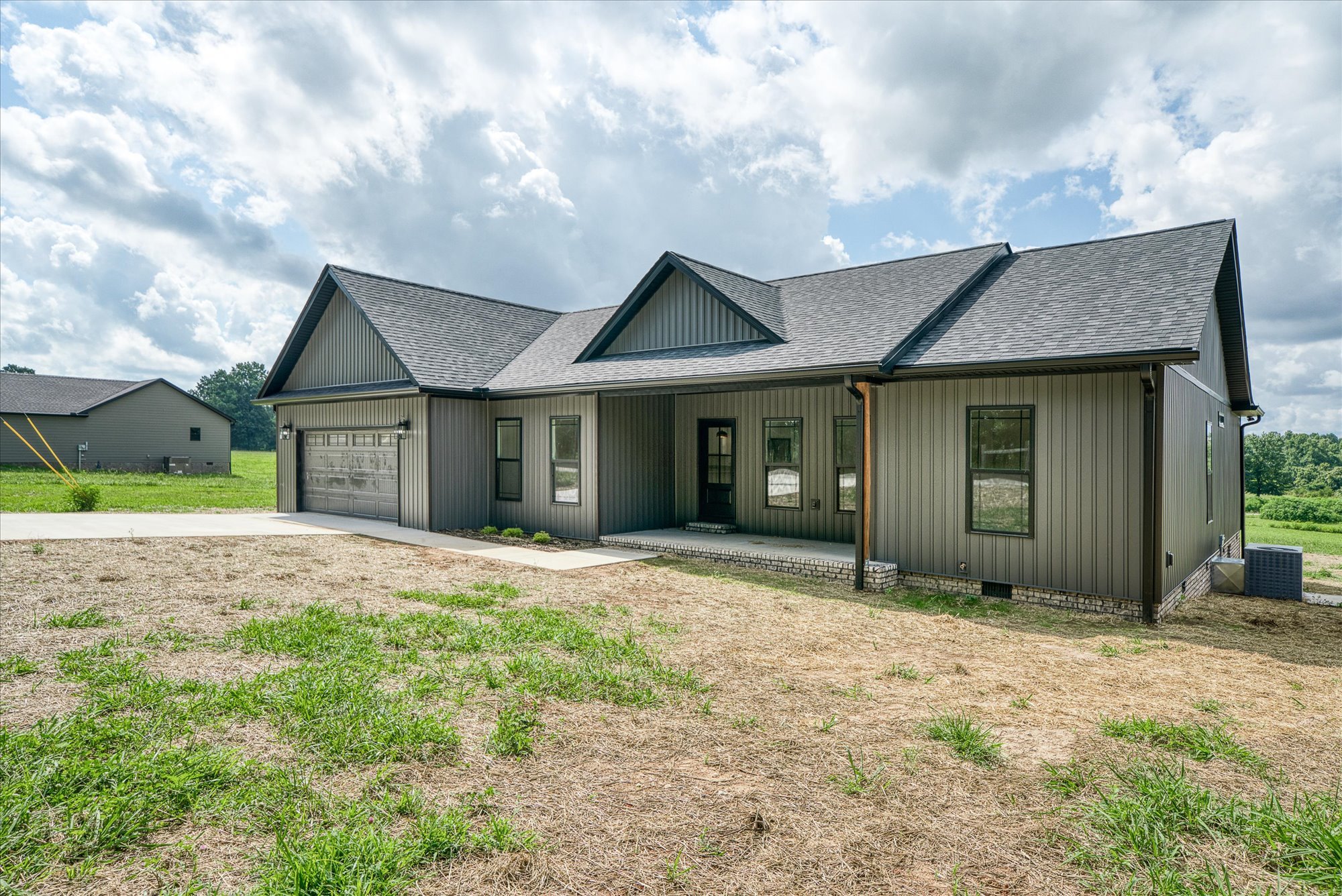 Front view of a modern house with a gray metal exterior and a gabled roof, located in a grassy area under a partly cloudy sky.