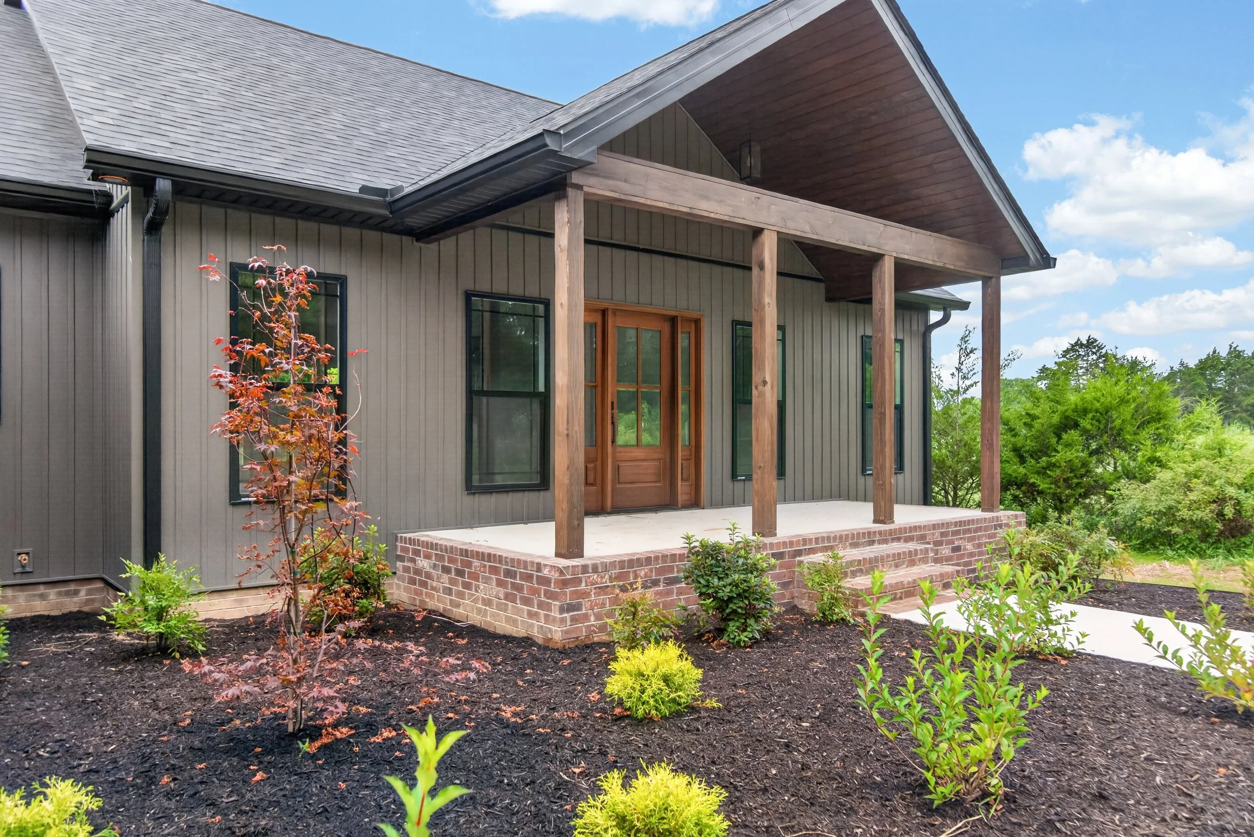 Front view of a house with a wooden porch, black siding, and brick steps, surrounded by a landscaped garden with young plants and trees under a blue sky with clouds.