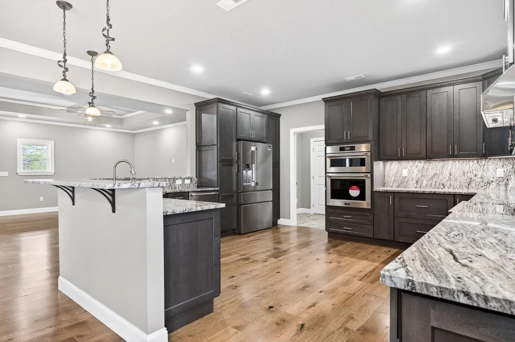 Modern kitchen with dark gray cabinets, granite countertops, hardwood floors, and stainless steel appliances, including a double oven and refrigerator.
