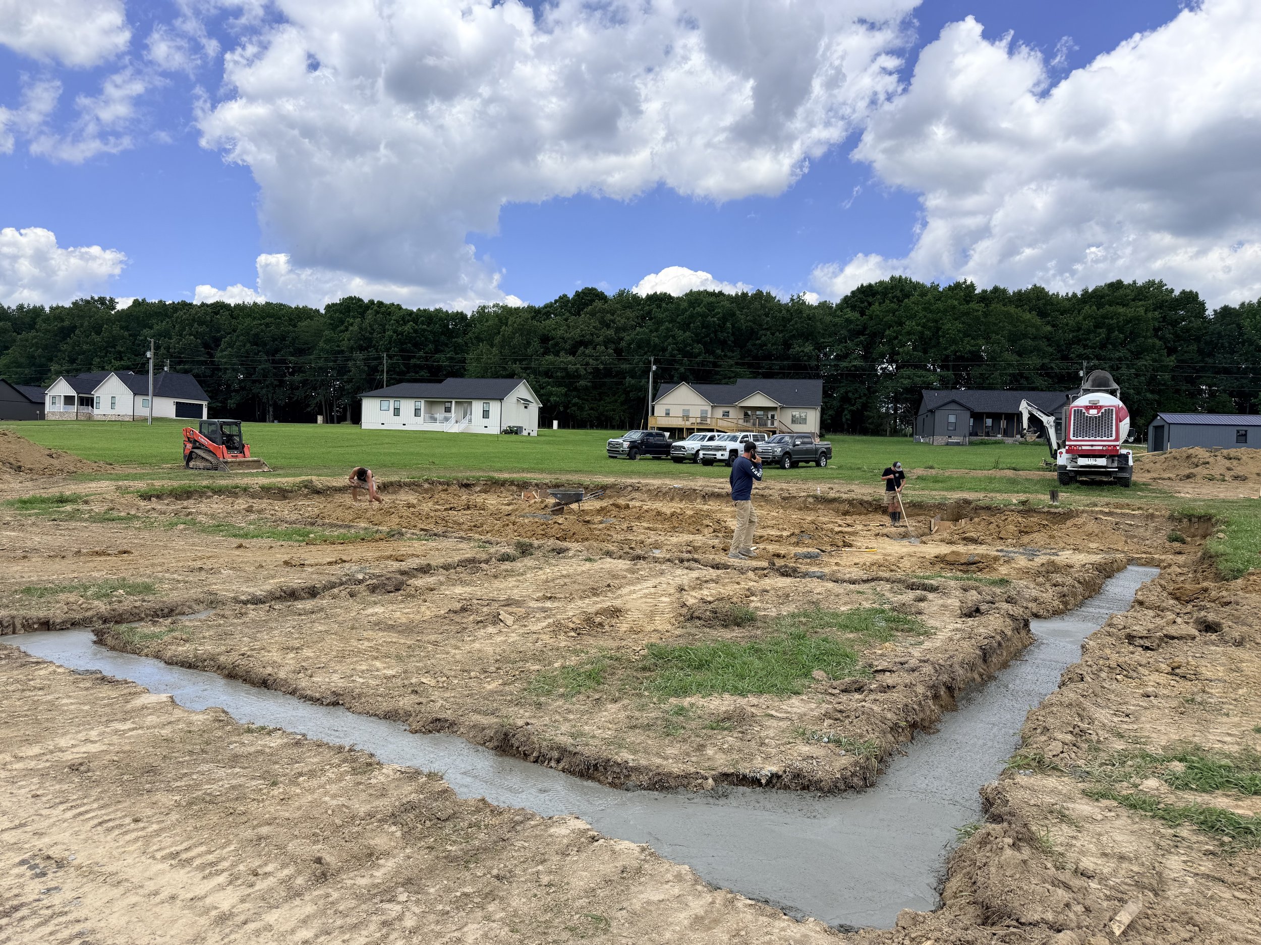 Construction site with workers and machinery in a grassy area, houses and trees in the background, and a partly cloudy sky overhead.