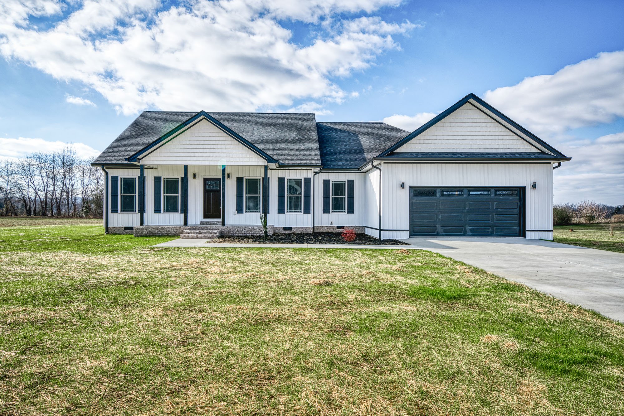 Front view of a white house with black shutters, and a black garage door, set on a grassy yard with a concrete driveway, under a partly cloudy sky.