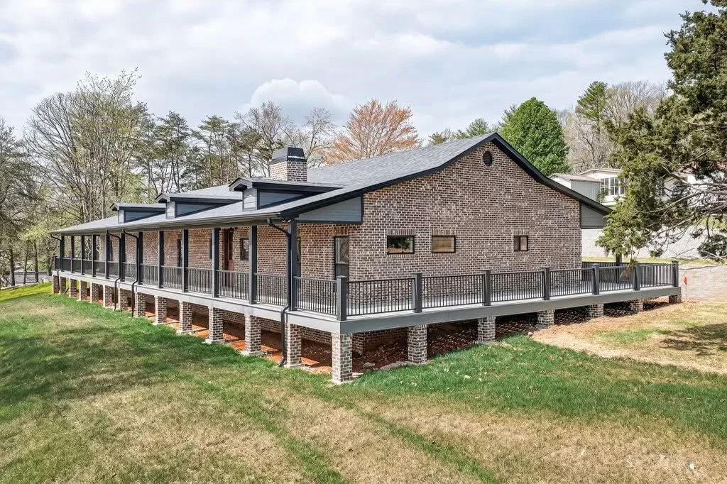A brick house with a large elevated porch surrounded by grass and trees.