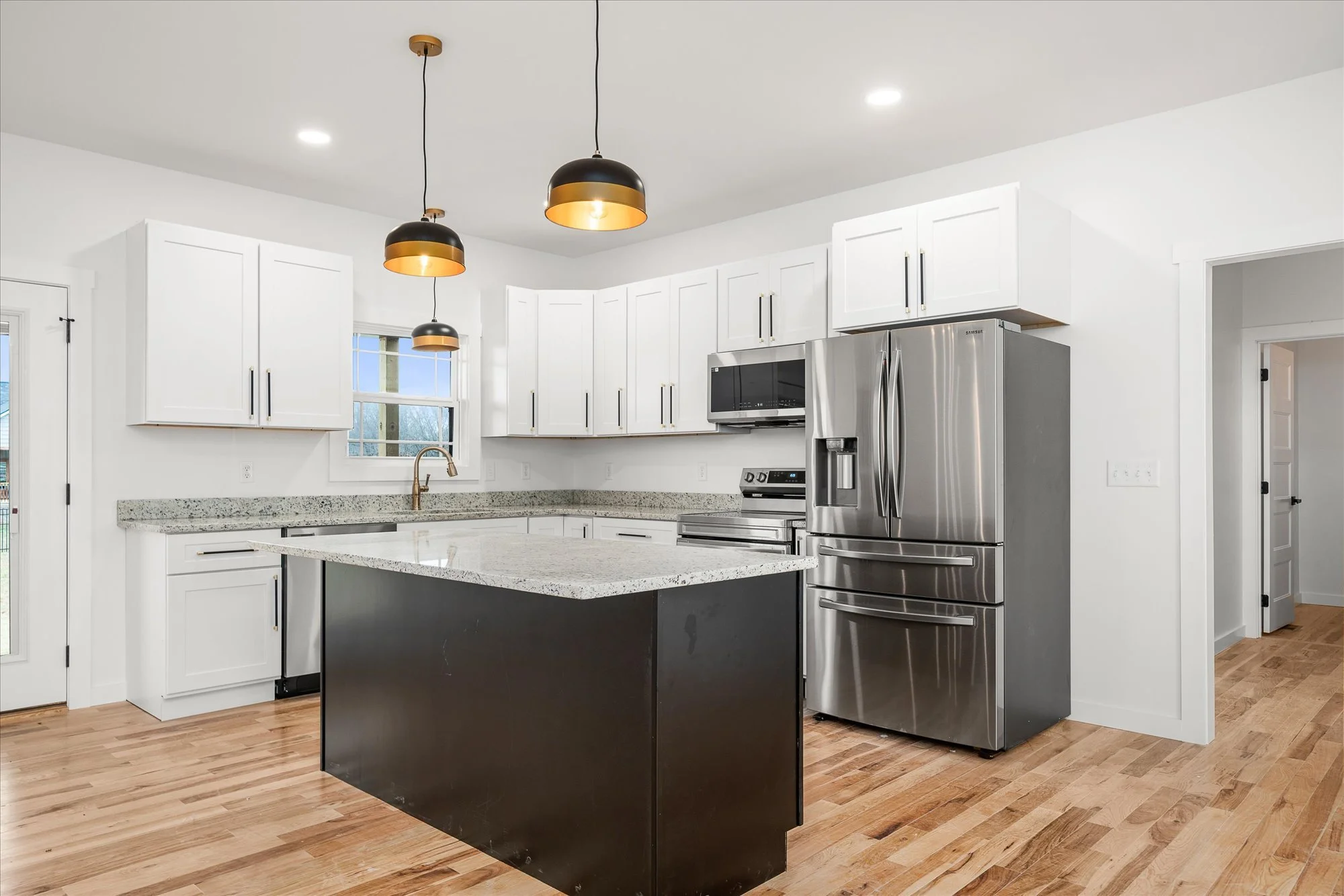 Modern kitchen with white cabinets, black and gold pendant lights, stainless steel appliances, a granite countertop island, and wood flooring.