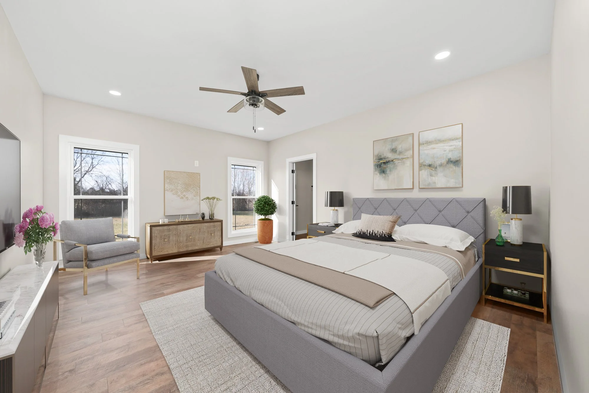 Bright bedroom with a bed, nightstands, lamps, artwork, windows, a ceiling fan, and a sitting area with a gray chair and flowers.