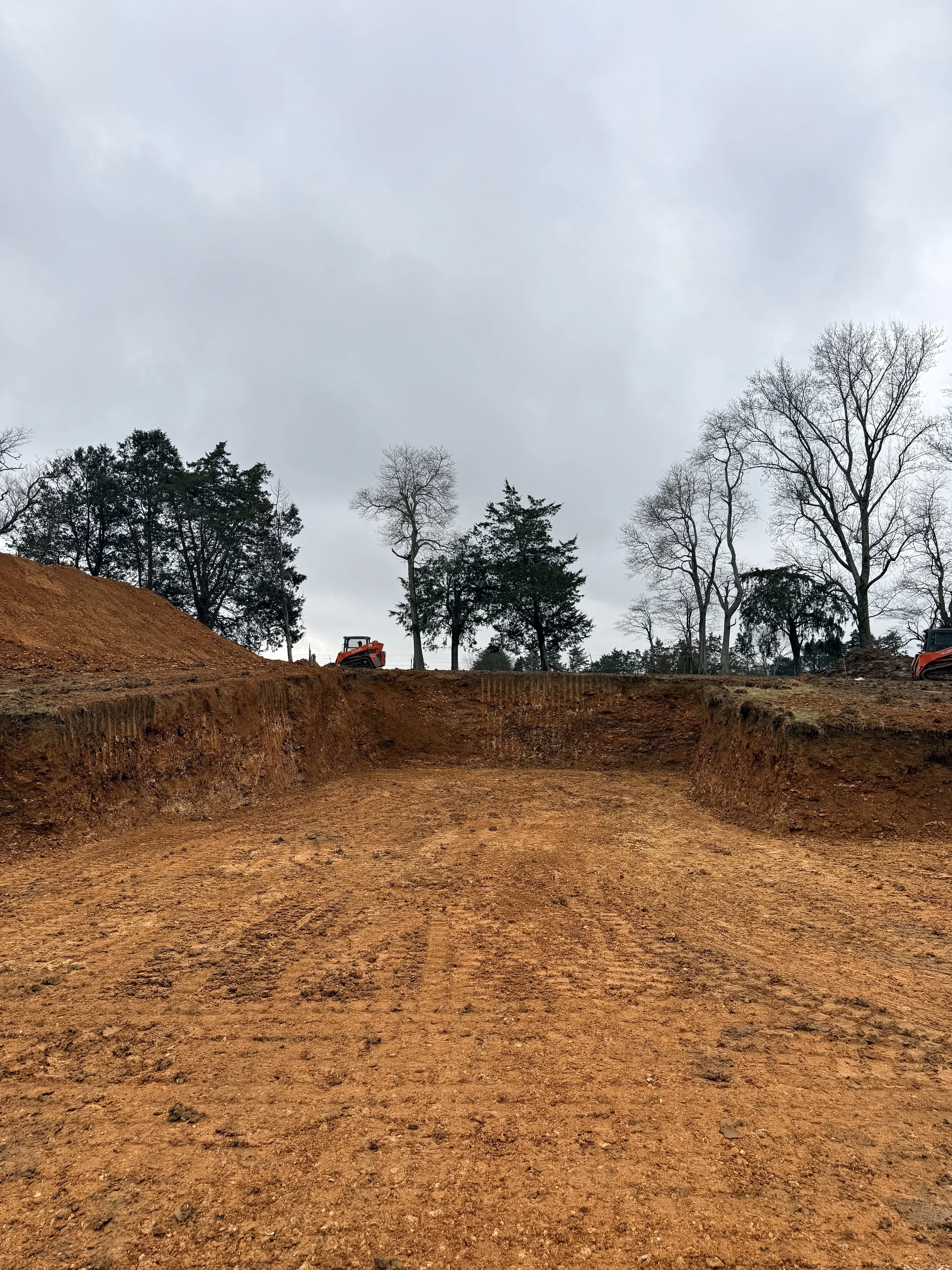 A construction site with freshly excavated dirt and a small orange excavator in the distance, under a cloudy sky with leafless trees.