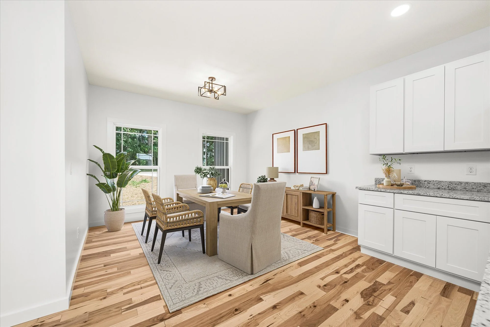 A bright, modern dining area with wooden flooring, a light-colored dining table surrounded by chairs, large windows providing natural light, and a decorative sideboard. There is a large potted plant near the window, and simple artwork on the walls. T