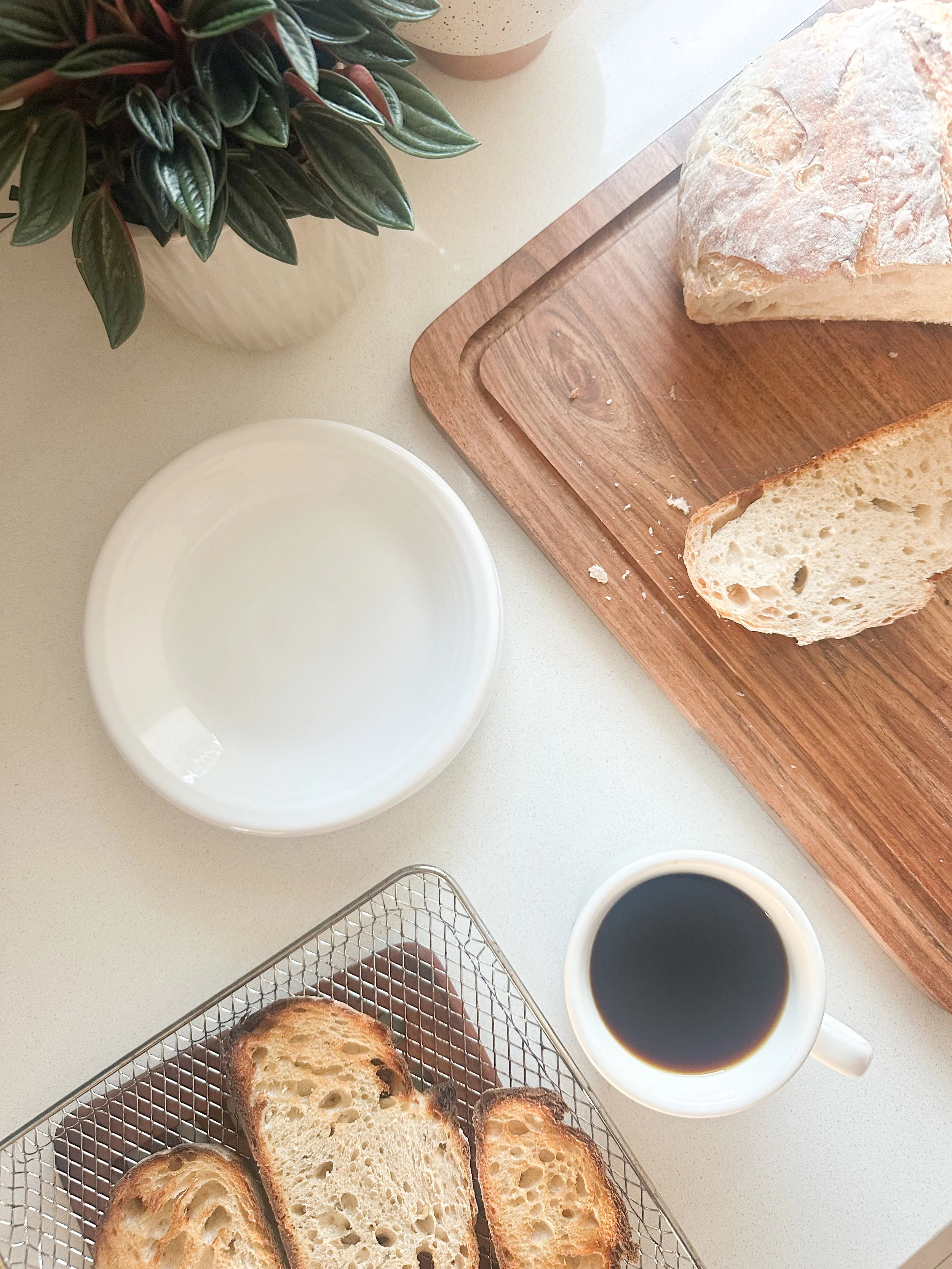 A photo showing slices of toasted bread in a wire basket, a loaf of bread on a wooden cutting board, a potted plant, a cup of black coffee, and an empty white bowl on a white countertop.
