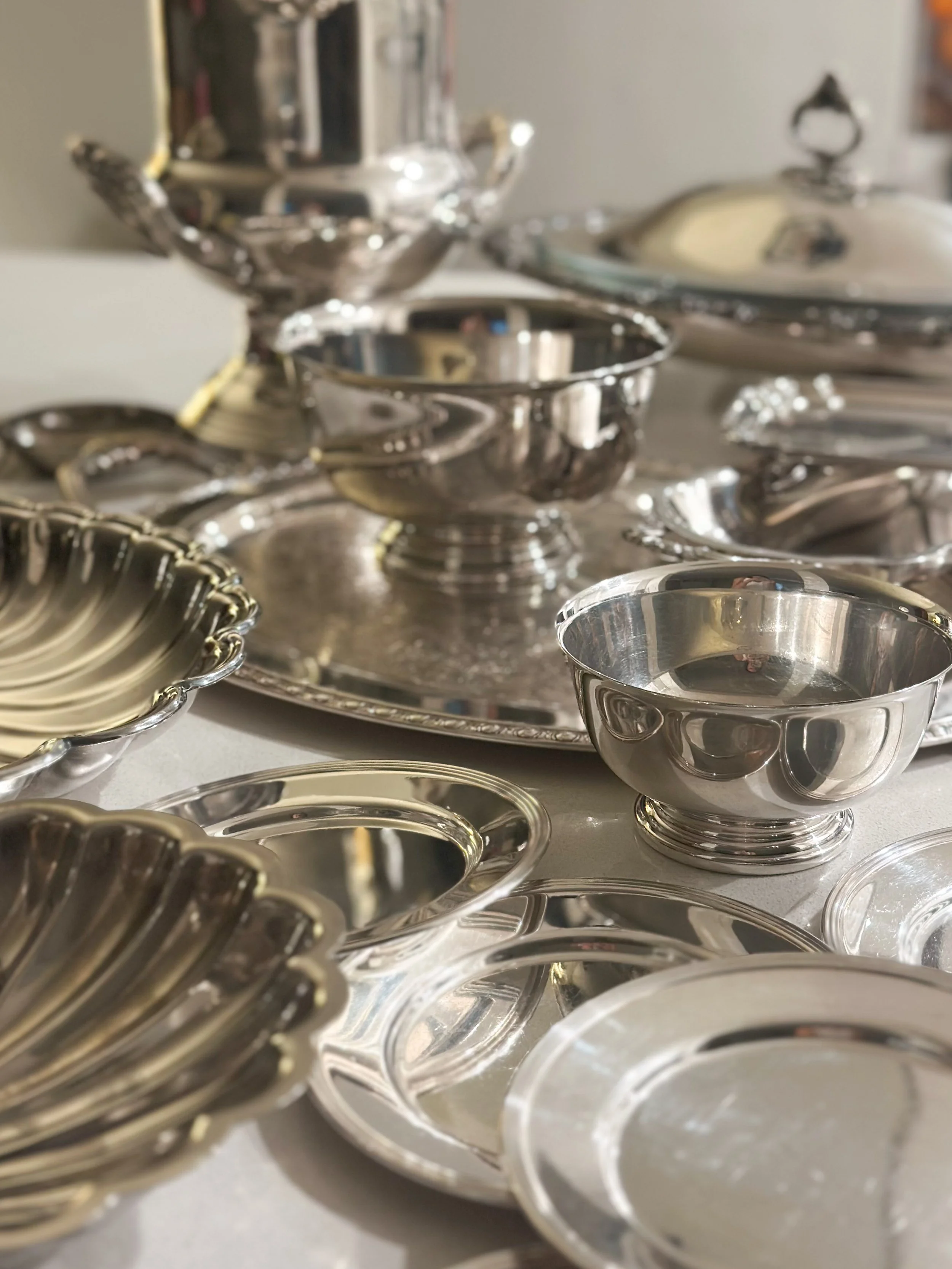 Multiple silver bowls and trays, some with scalloped edges, arranged on a flat surface. The background contains a large silver serving piece with a lid.