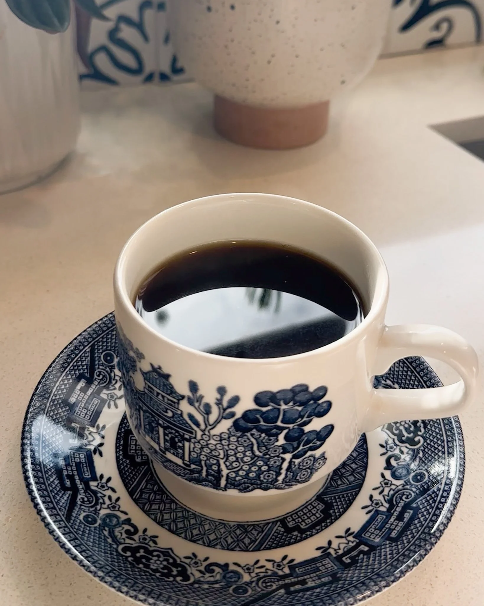 Close-up of a cup of black coffee on a decorative blue and white saucer with intricate patterns, placed on a light-colored surface. In the background, a blue and white blue willow mug and a decorative vases with a similar color scheme are visible.