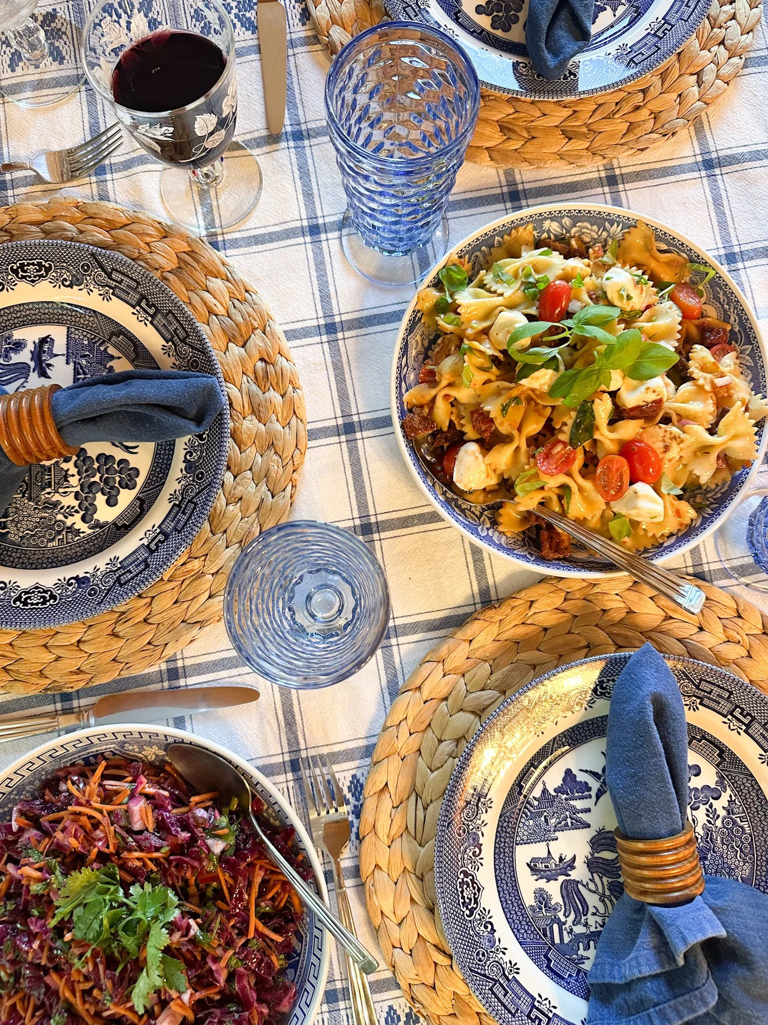 A table set for a meal with patterned plates, woven placemats, glassware, a bowl of pasta salad garnished with basil, a glass of red wine, and cutlery with blue cloth napkins.